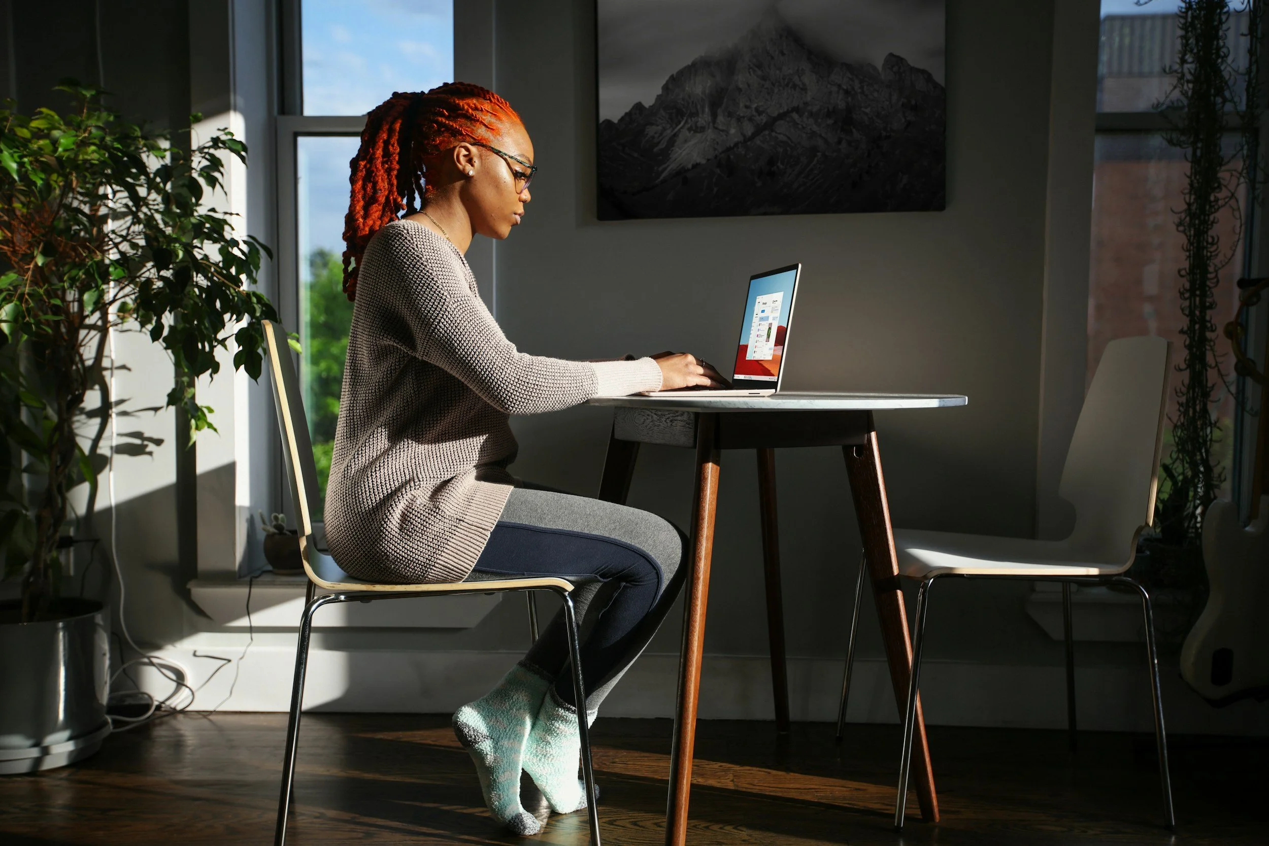 A woman with braided red hair, glasses, and a beige sweater working on a laptop at a wooden dining table in a sunlit room with plants and a landscape painting on the wall.