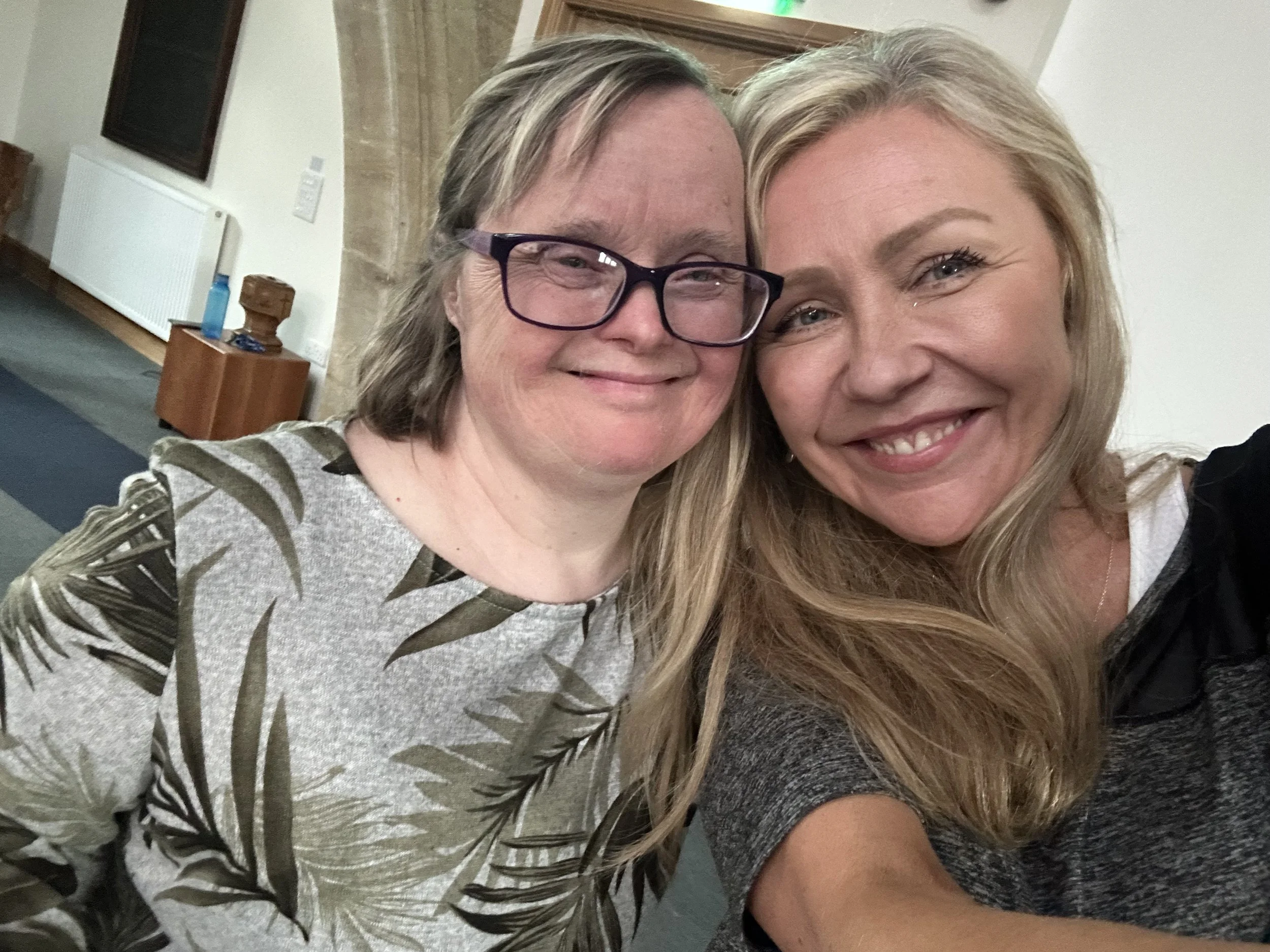 Two women taking a close-up selfie together, smiling and looking happy. The woman on the left has glasses and shoulder-length hair, wearing a top with a leafy pattern. The woman on the right has long blonde hair and is wearing a dark top.