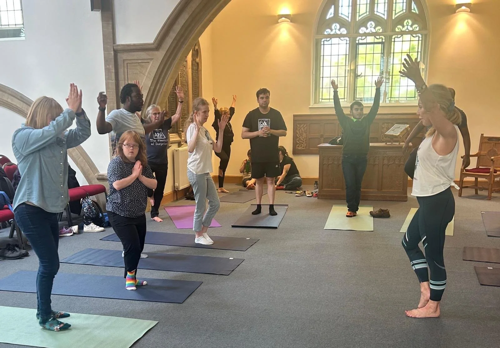 People participating in a yoga class inside a church or chapel, with large stained glass windows, some participants standing on yoga mats with hands raised or in prayer positions.