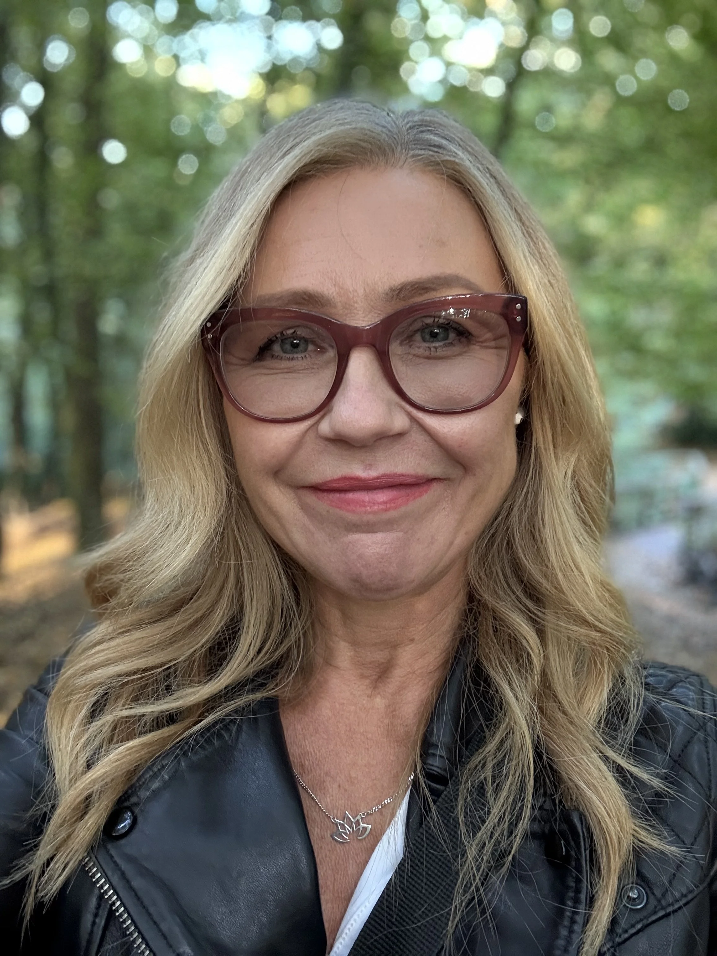 Jane Wrafter Muswell Hill Therapy with long blonde hair wearing large red glasses and a black leather jacket, outdoors in a wooded area with green trees in the background.