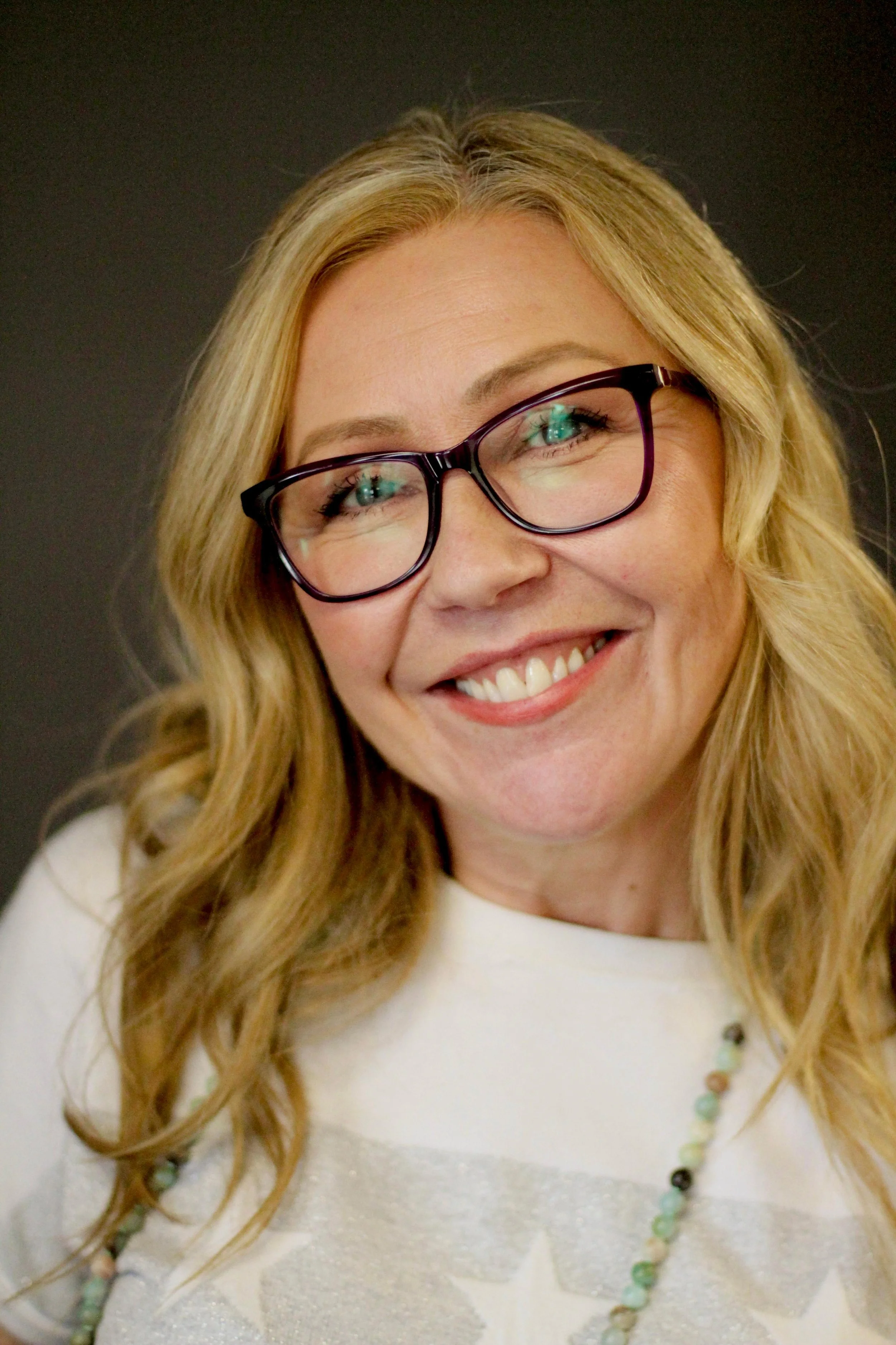 Jane Wrafter Muswell Hill Therapy with long blonde wavy hair, glasses, and a big smile, wearing a white top and a multicolored beaded necklace, against a dark background.