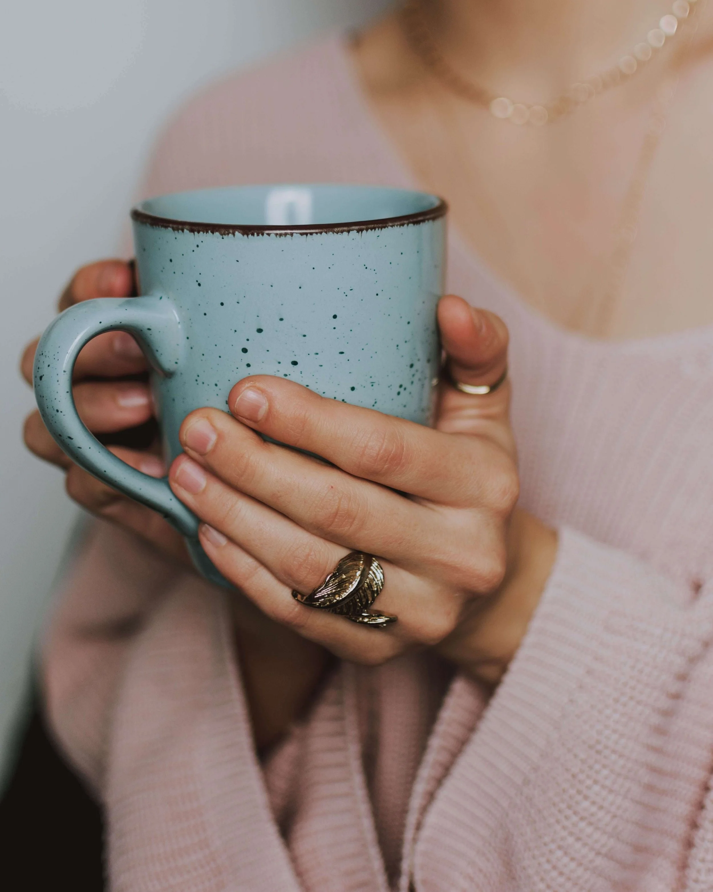 Person holding a large speckled blue ceramic mug, wearing a pink sweater and a pearl necklace, with a gold ring on their right hand.