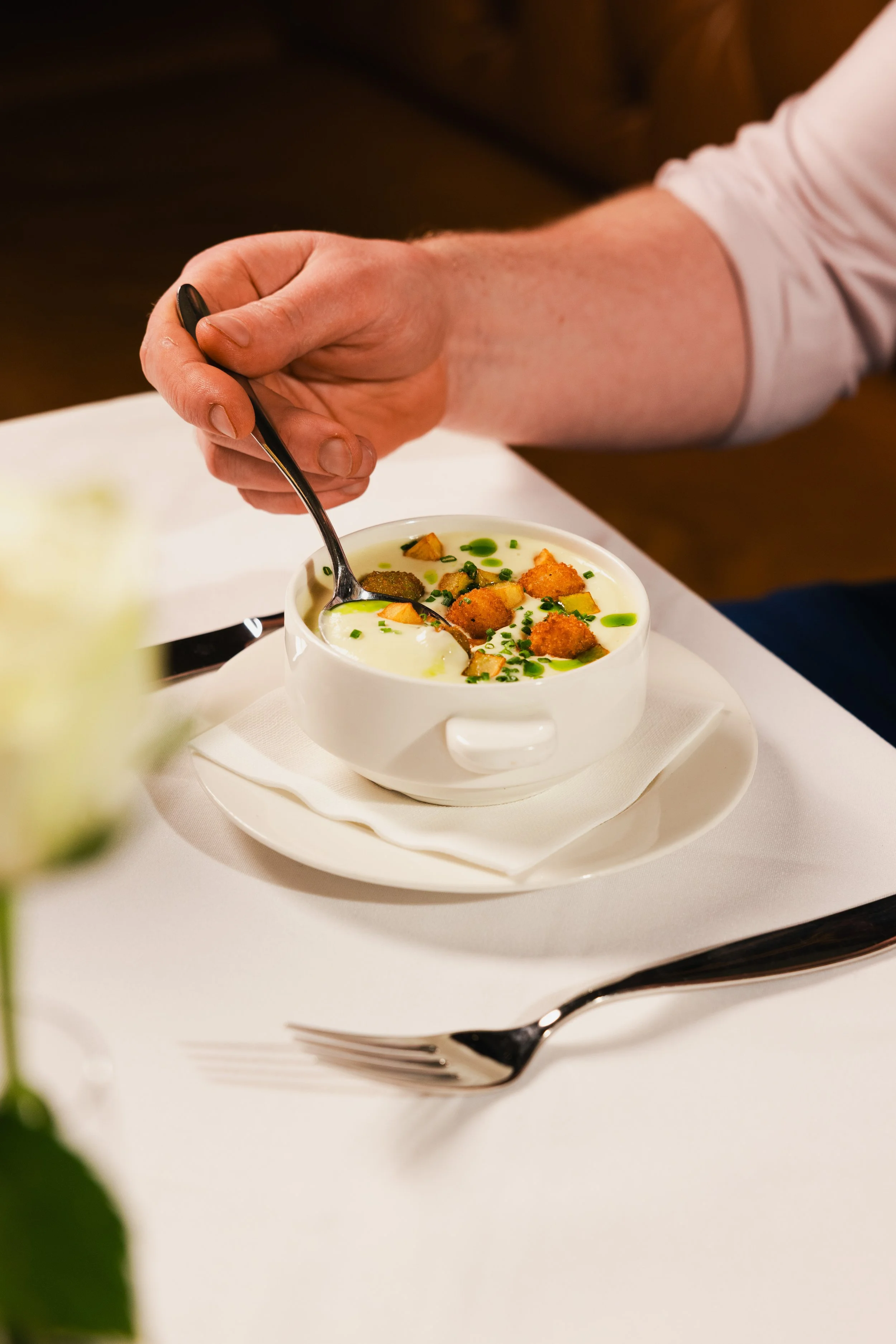 A person is using a spoon to eat soup from a white bowl on a white tablecloth. The soup is garnished with chopped chives, croutons, and a drizzle of green oil. A fork is placed beside the bowl, and a blurred green flower is visible in the foreground.