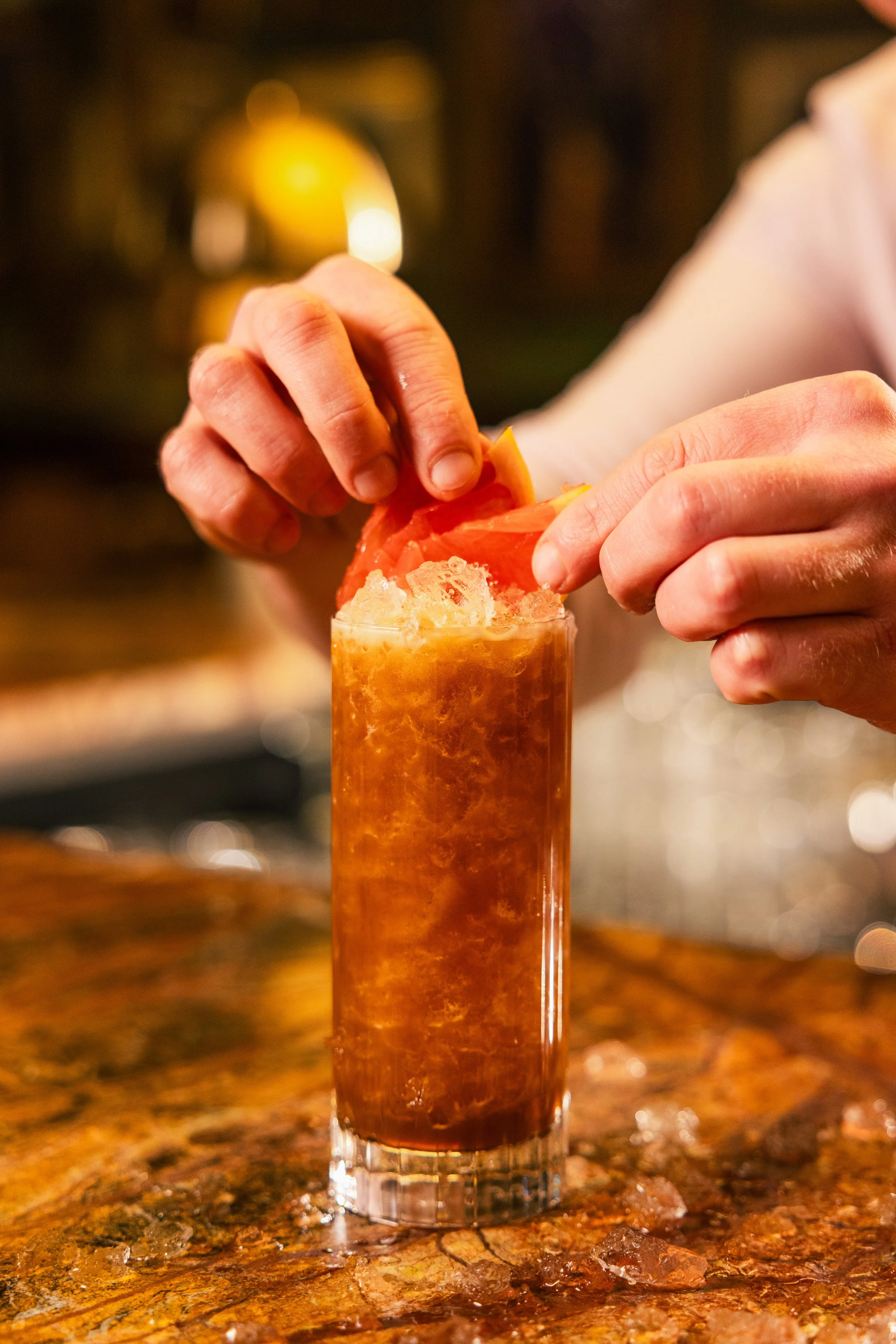 Close-up of a person filling a tall glass with a dark cocktail and ice, with a blurred warm-lit background.