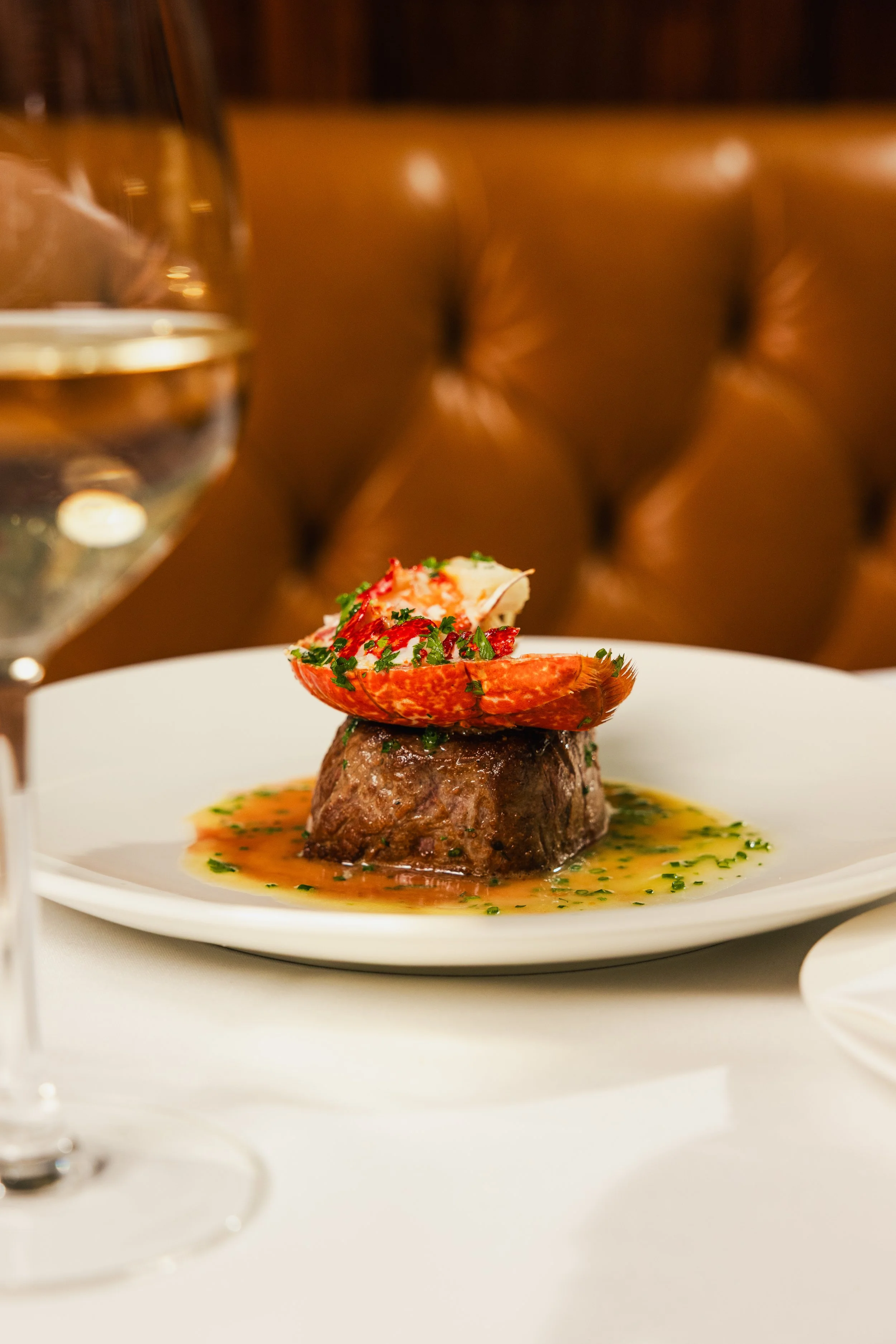 A plated gourmet dish featuring a meat centerpiece topped with seafood and herbs, garnished with a sauce and served in an elegant restaurant setting with a glass of water nearby.