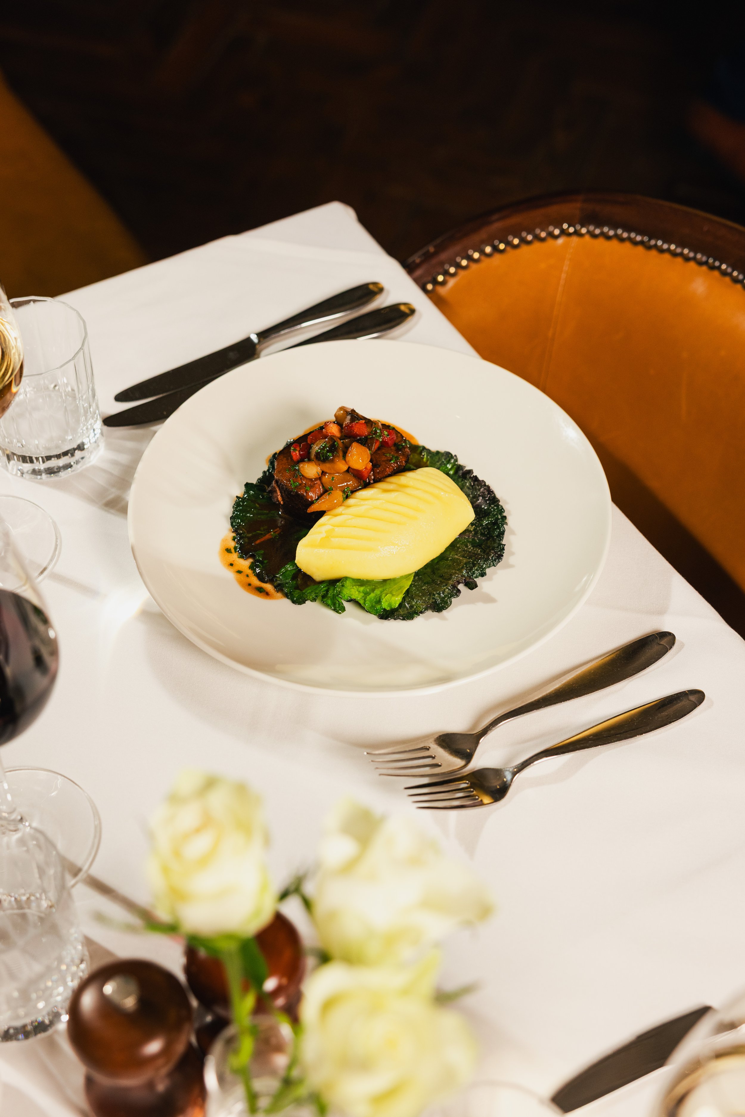 A plated gourmet meal with a piece of meat topped with vegetables, mashed potatoes, and leafy greens on a white plate, set on a white tablecloth with silverware.
