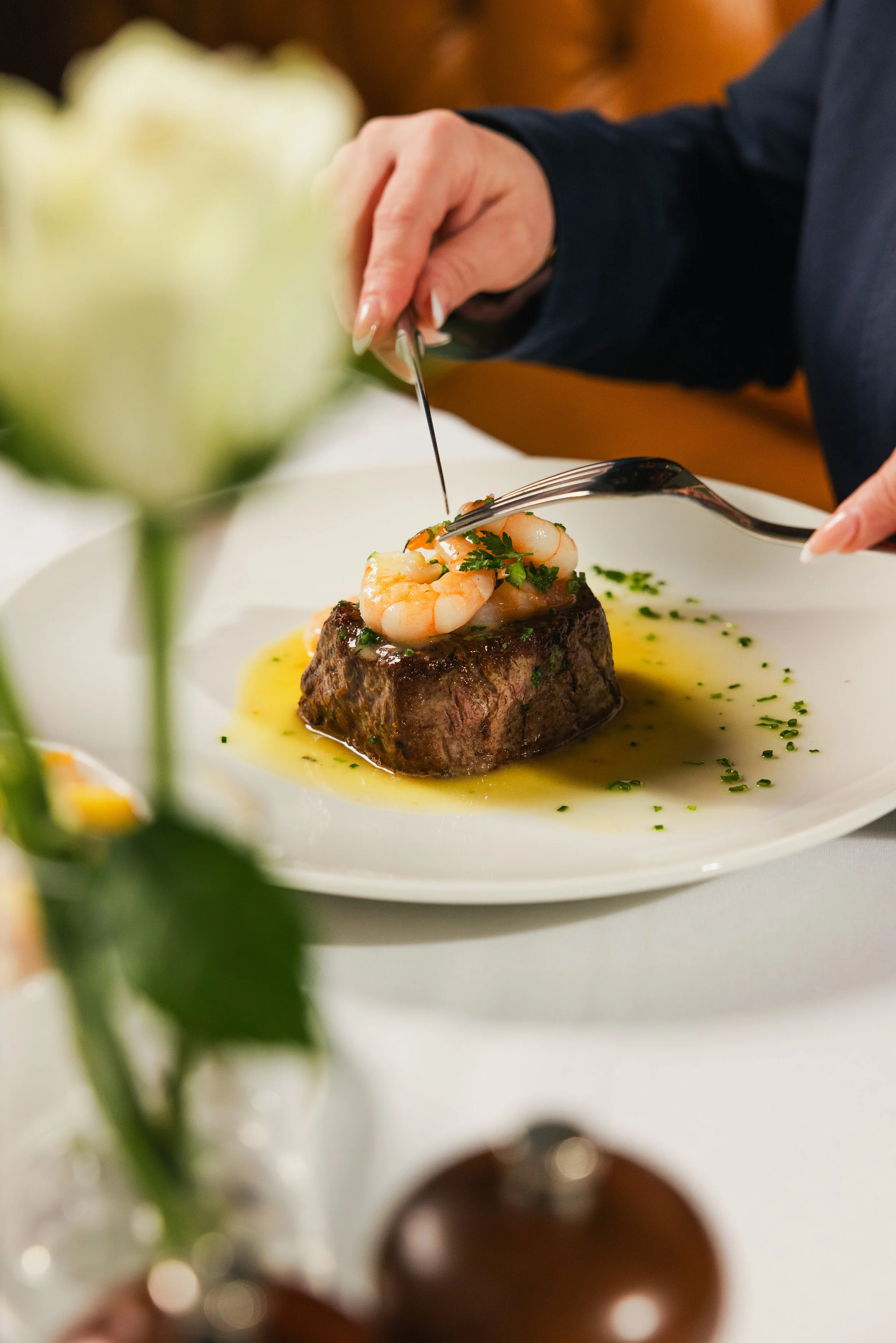 Close-up of a plated gourmet dish featuring a seared steak topped with shrimp and garnished with herbs, drizzled with sauce, and presented on a white plate with a decorative flower in the foreground.