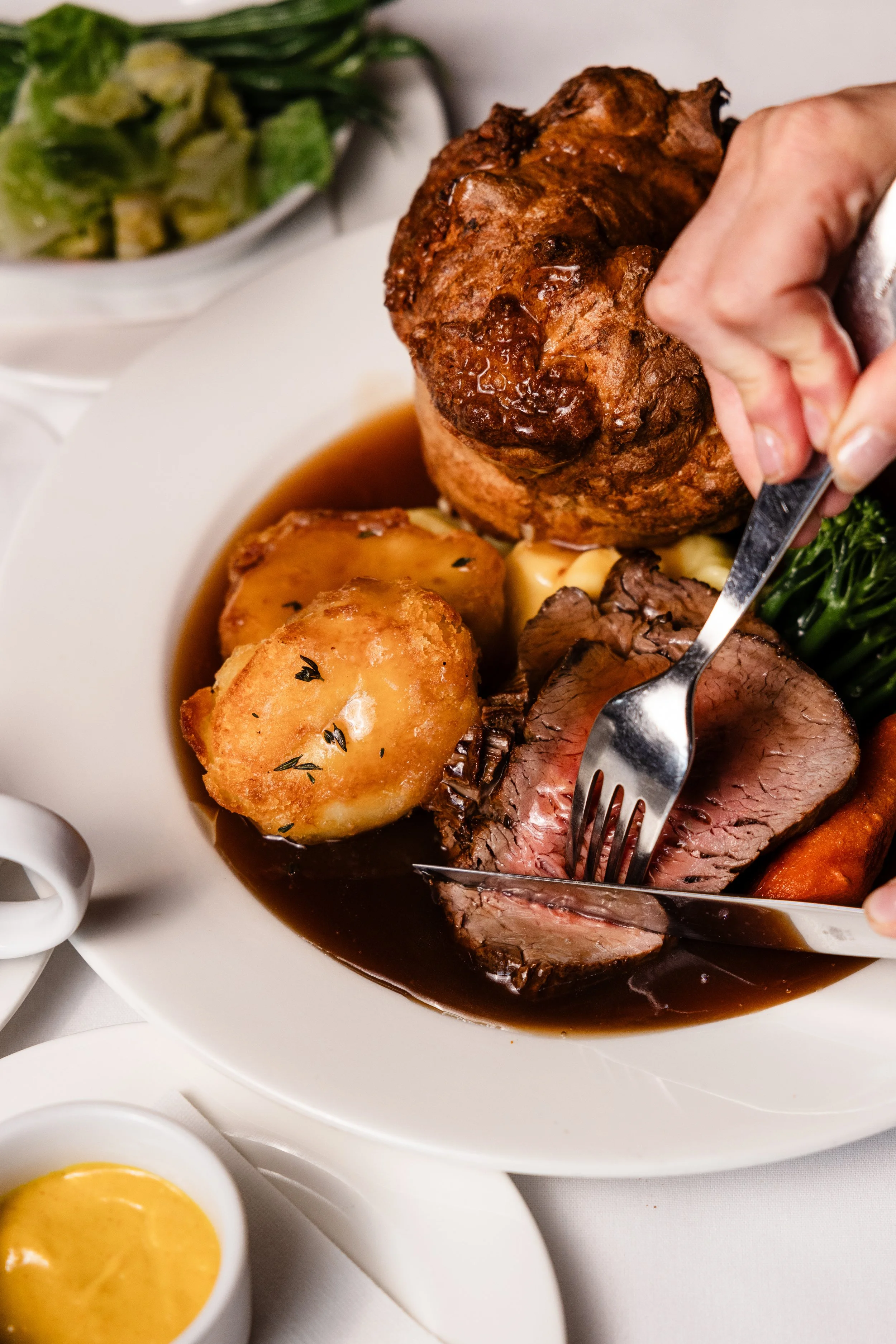 A plate of roast beef with juice, roasted potatoes with herbs, gravy, and green vegetables, with small bowls of yellow sauce on the side.