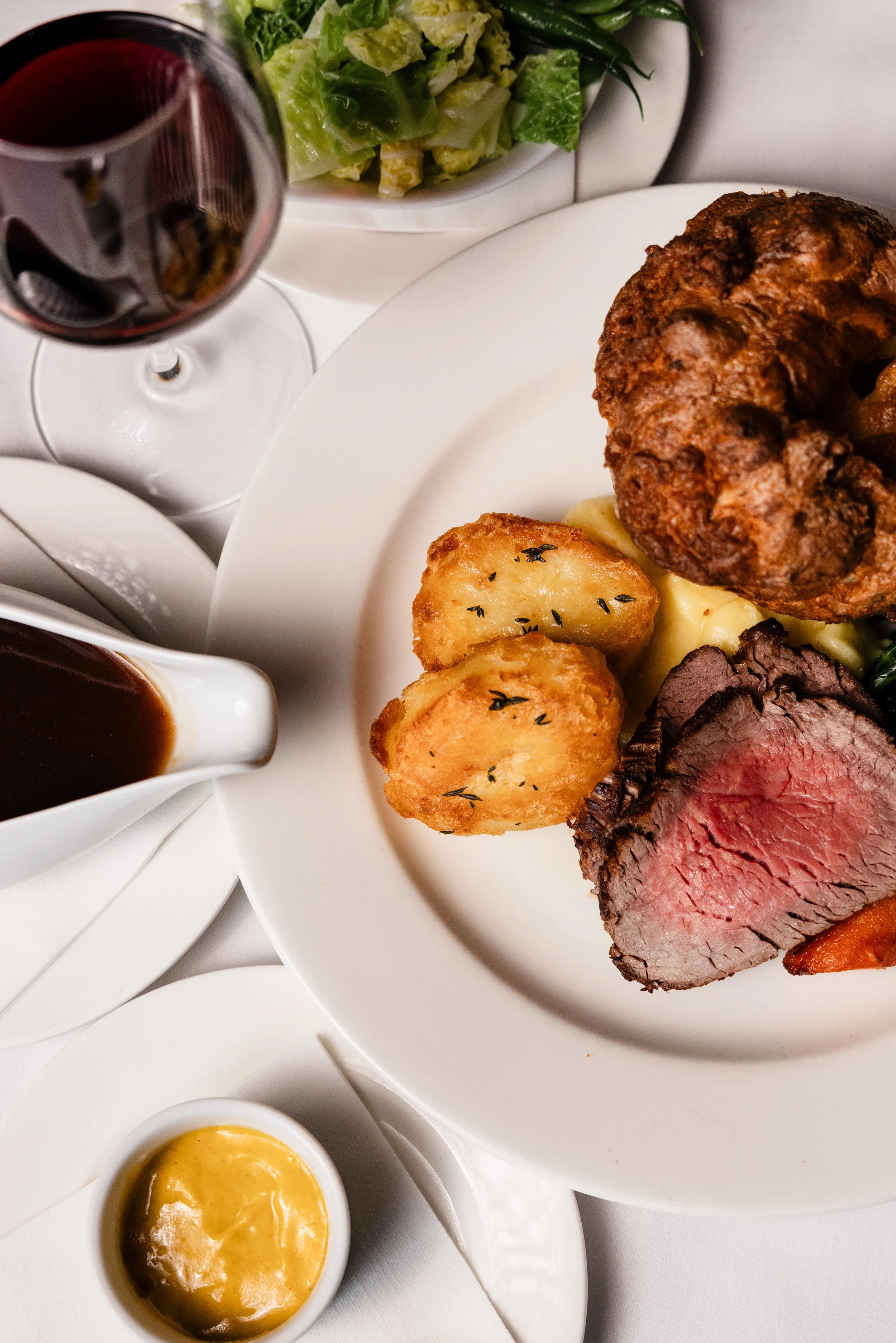 A plate of roast beef with golden roasted potatoes garnished with herbs, served with gravy. A side of mixed green salad and green beans is visible in the background. A glass of red wine and a small bowl of mustard are also present.