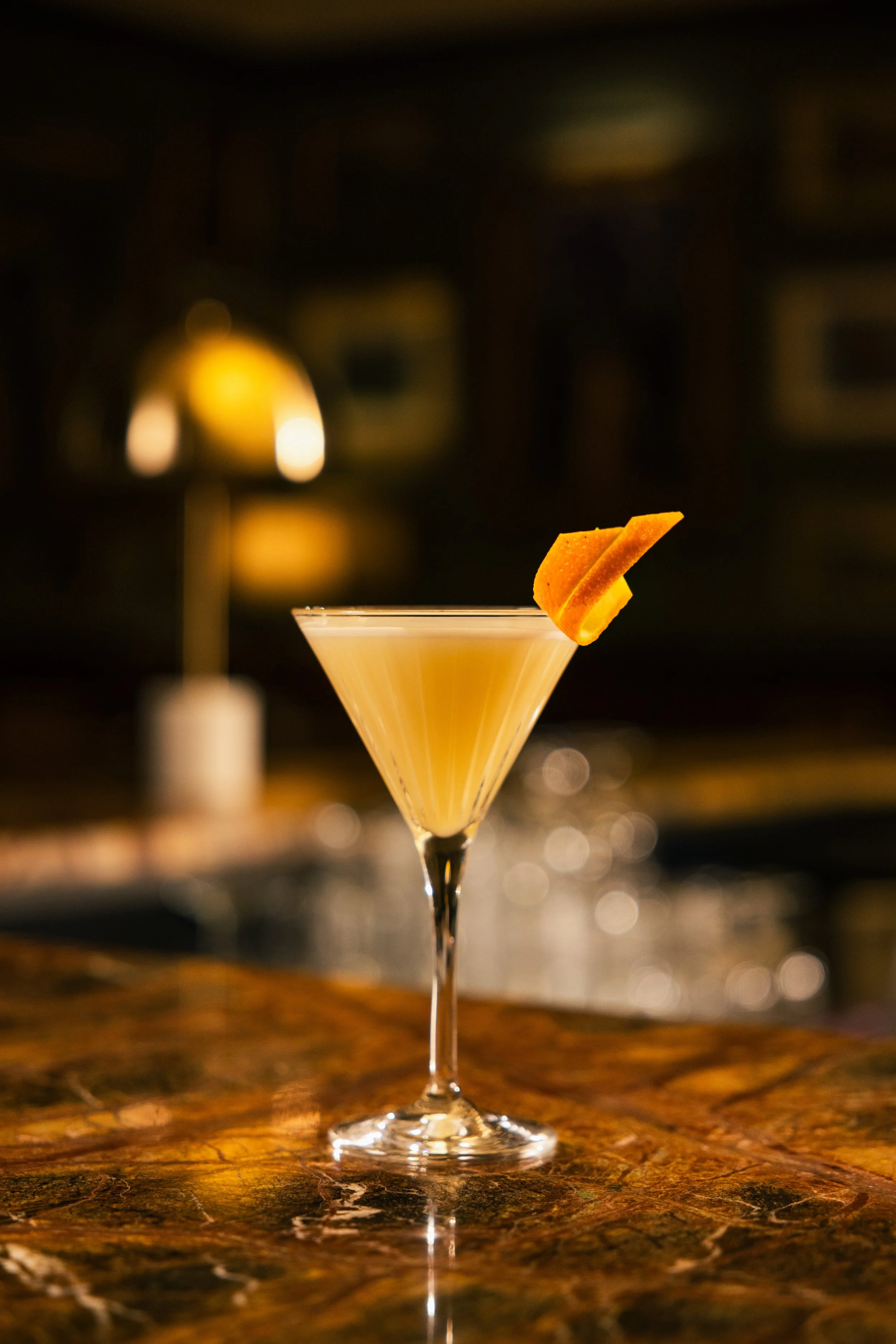 A cocktail glass with a lemon peel garnish on the rim, set on a polished dark marble bar in a dimly lit bar or restaurant.