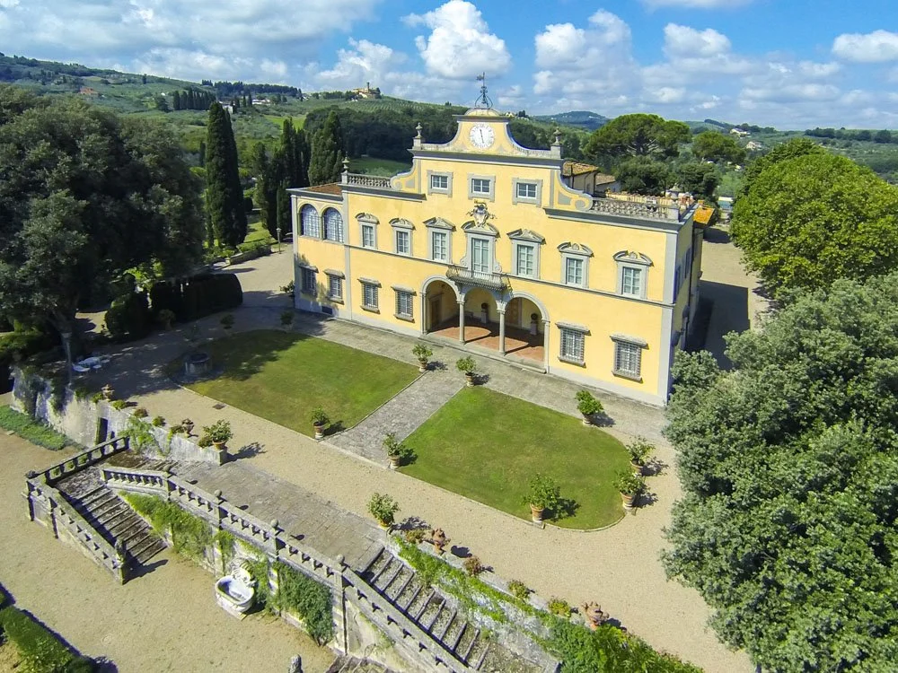 A yellow historic villa with a clock on top, surrounded by lush greenery and a landscaped garden, situated in a hilly countryside.