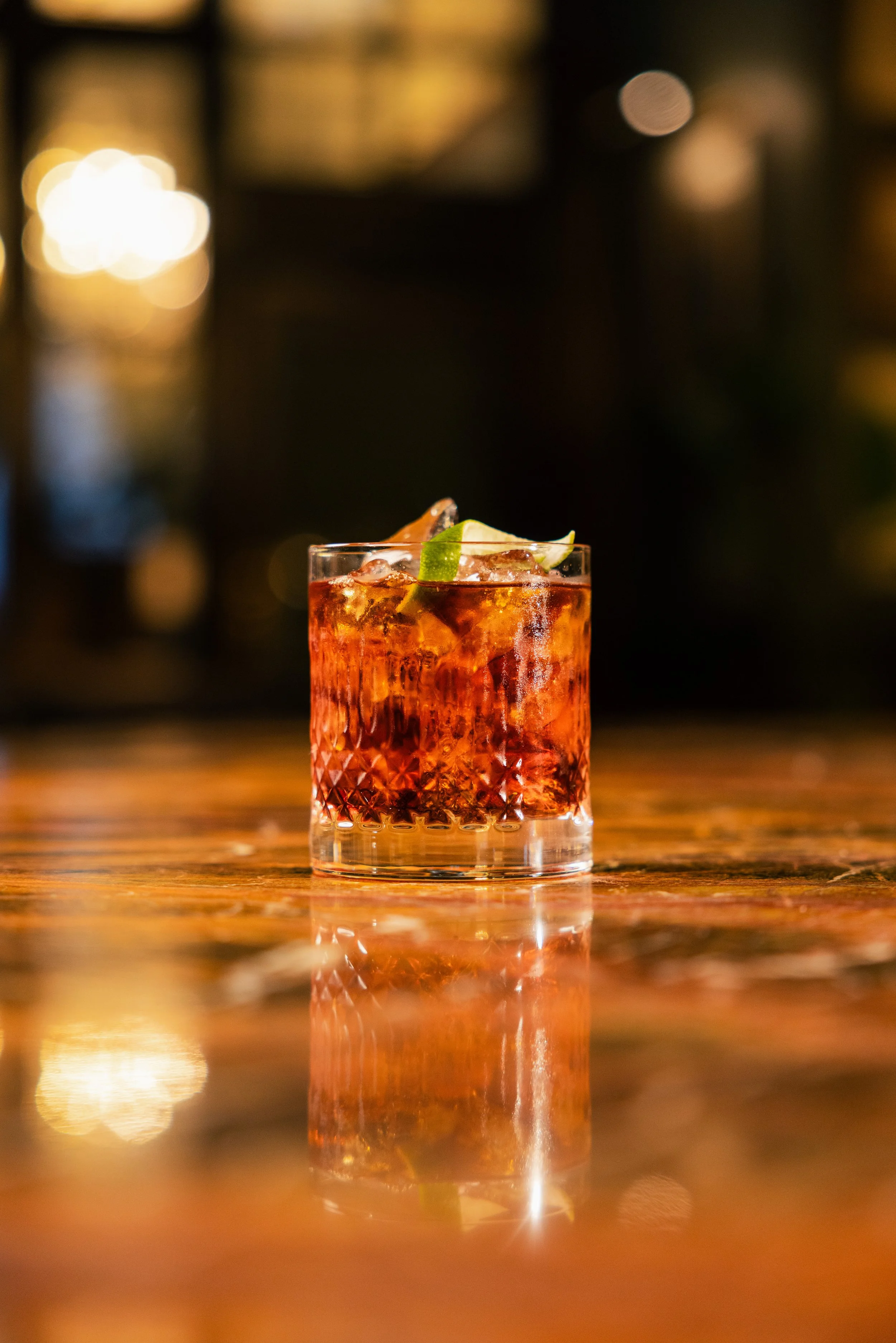 A glass of red cocktail garnished with lime slices on a wooden table, with blurred warm lights in the background.