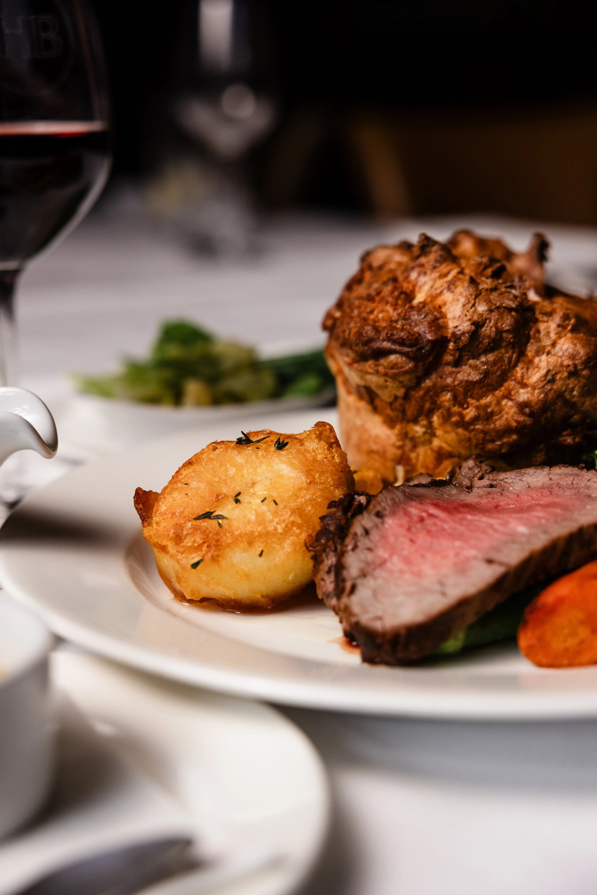Close-up of a plated meal with roasted meat, a baked potato garnished with herbs, and green vegetables, with a glass of red wine on a white tablecloth.