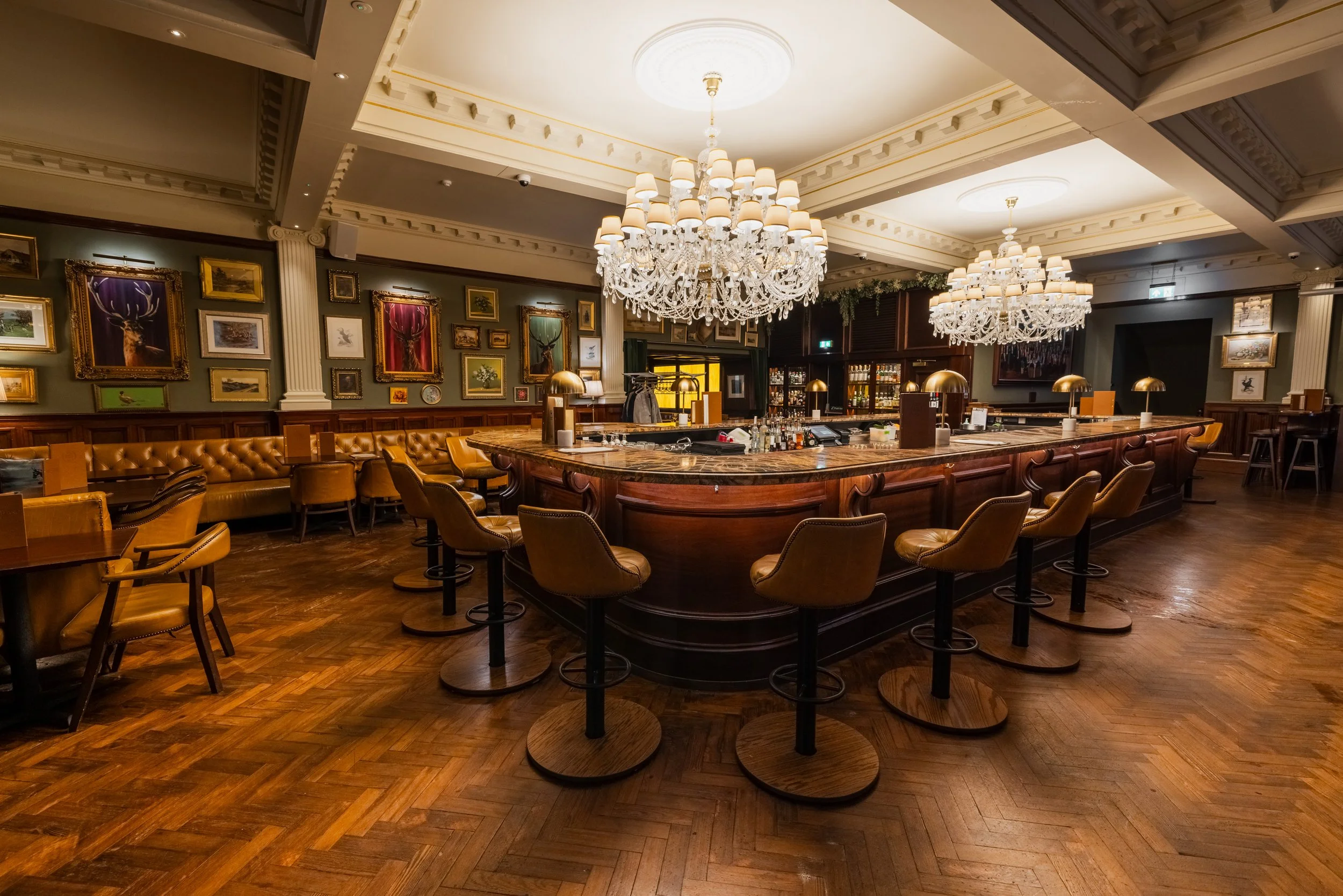 Elegant bar with large crystal chandeliers, wooden bar counter, leather chairs, yellow wood flooring, and framed artwork on the walls.
