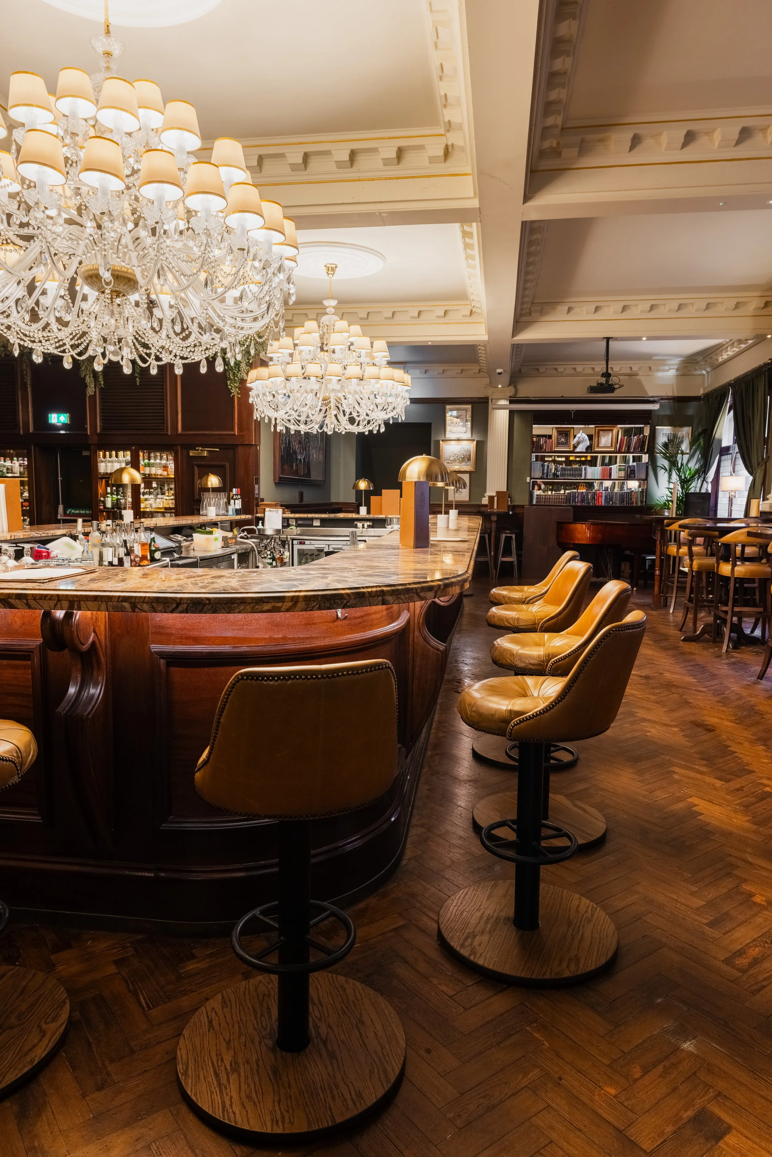 Interior of a luxurious bar or lounge with a marble-topped curved bar counter, high leather stools with circular wooden bases, several chandeliers with lampshades, dark wood furnishings, a bookshelf filled with books, and large windows with curtains.