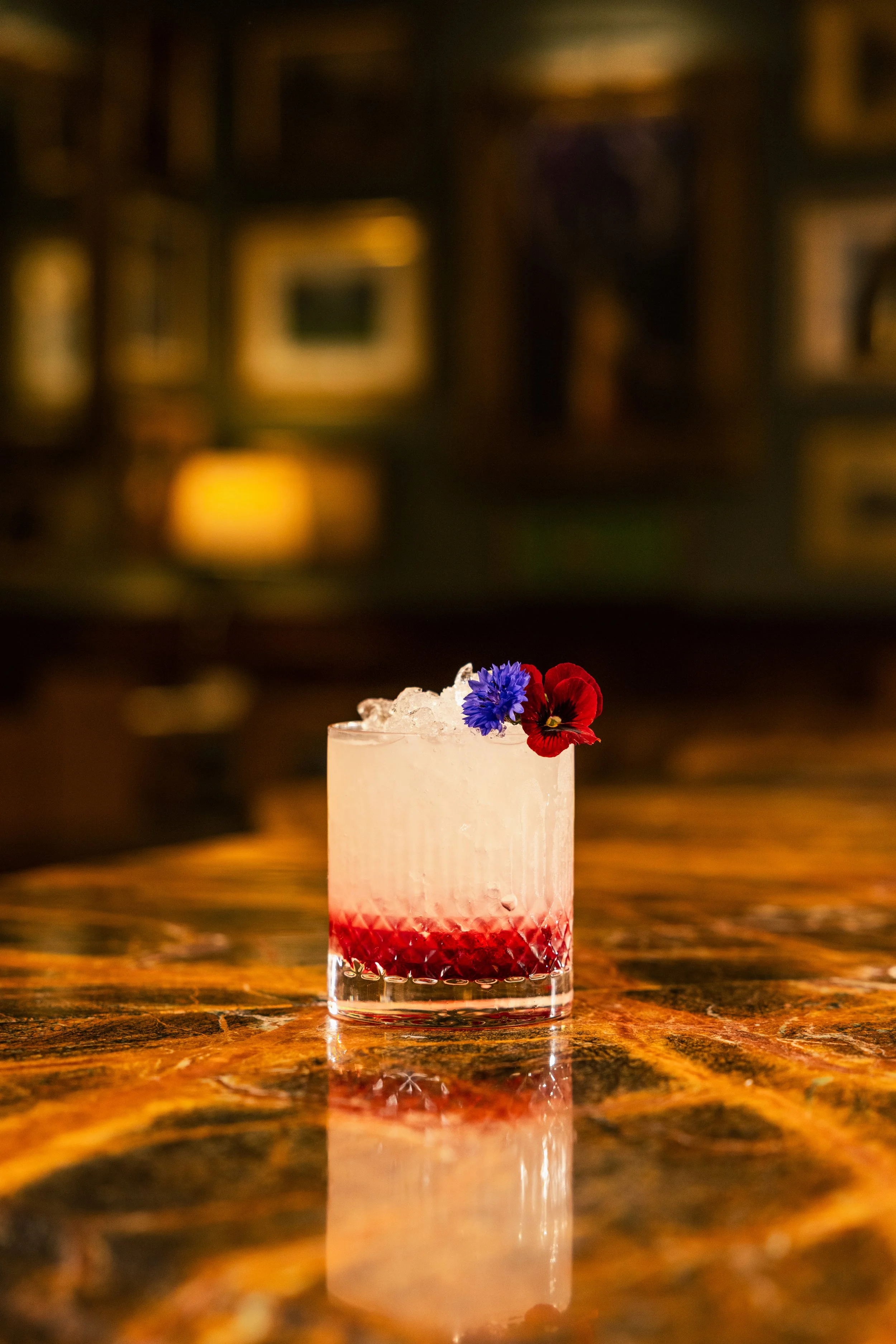 A cocktail in a short glass with red layered fruit at the bottom, topped with crushed ice, garnished with a blue cornflower and a red pansy flower, on a wooden table.