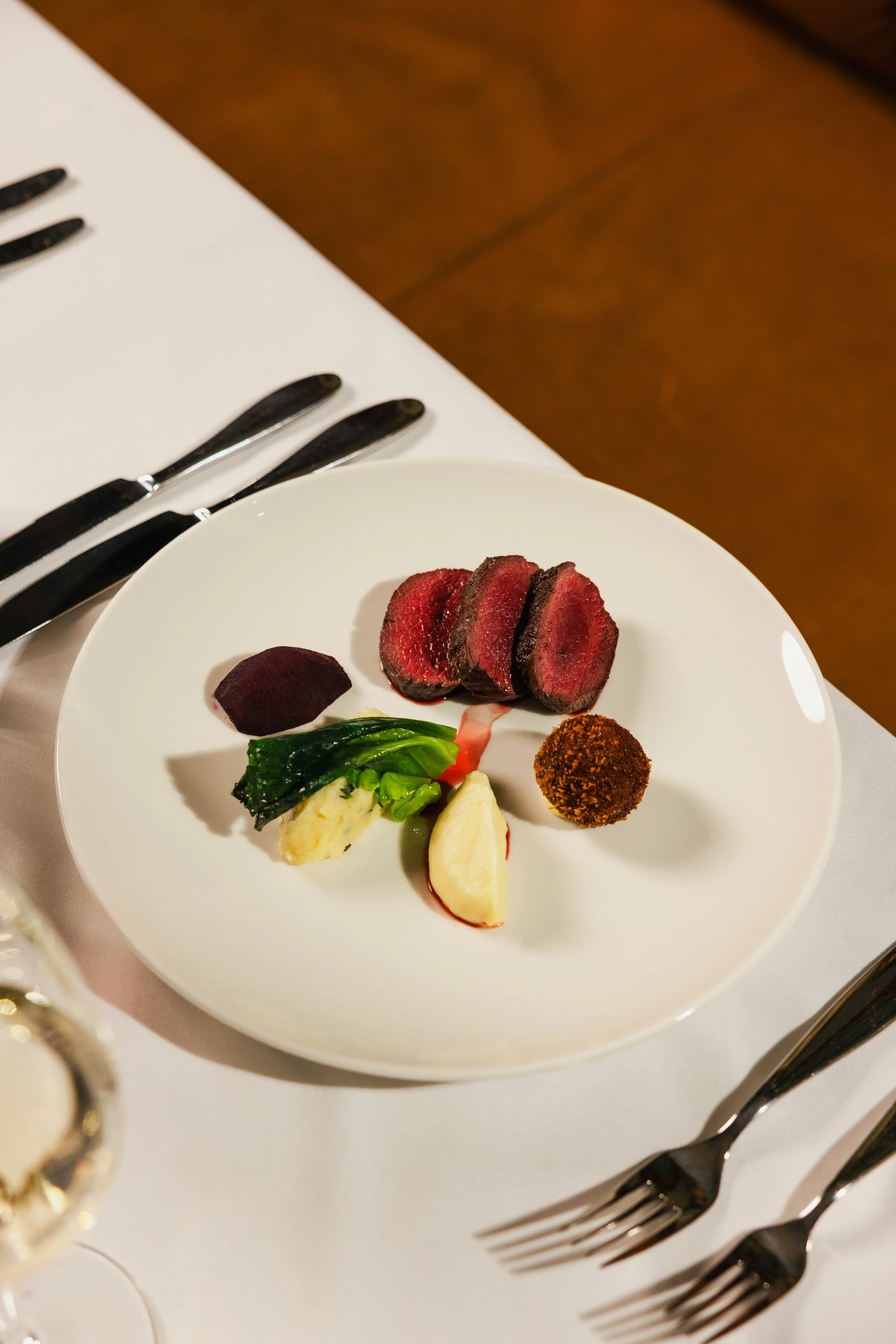 A white plate with sliced medium-rare steak, a beet slice, cooked greens, pieces of potato, and a small ball of breaded food, set on a table with silverware and a white tablecloth.