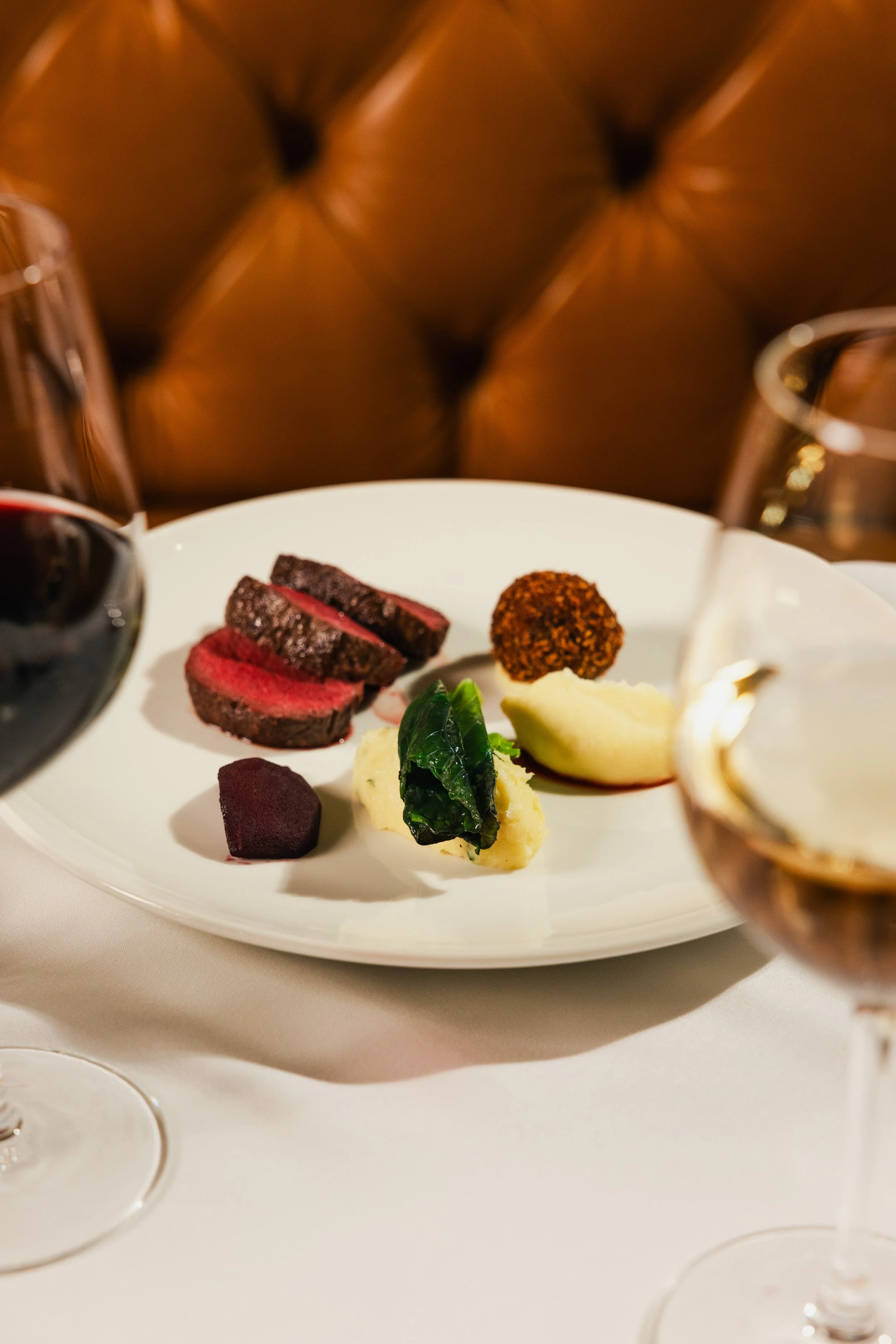 A white plate with slices of cooked meat, a round fried item, mashed potatoes with a green leafy garnish, and some deep purple vegetables. There are two glasses of red wine partially visible on either side of the plate, and the background features a brown tufted headboard.