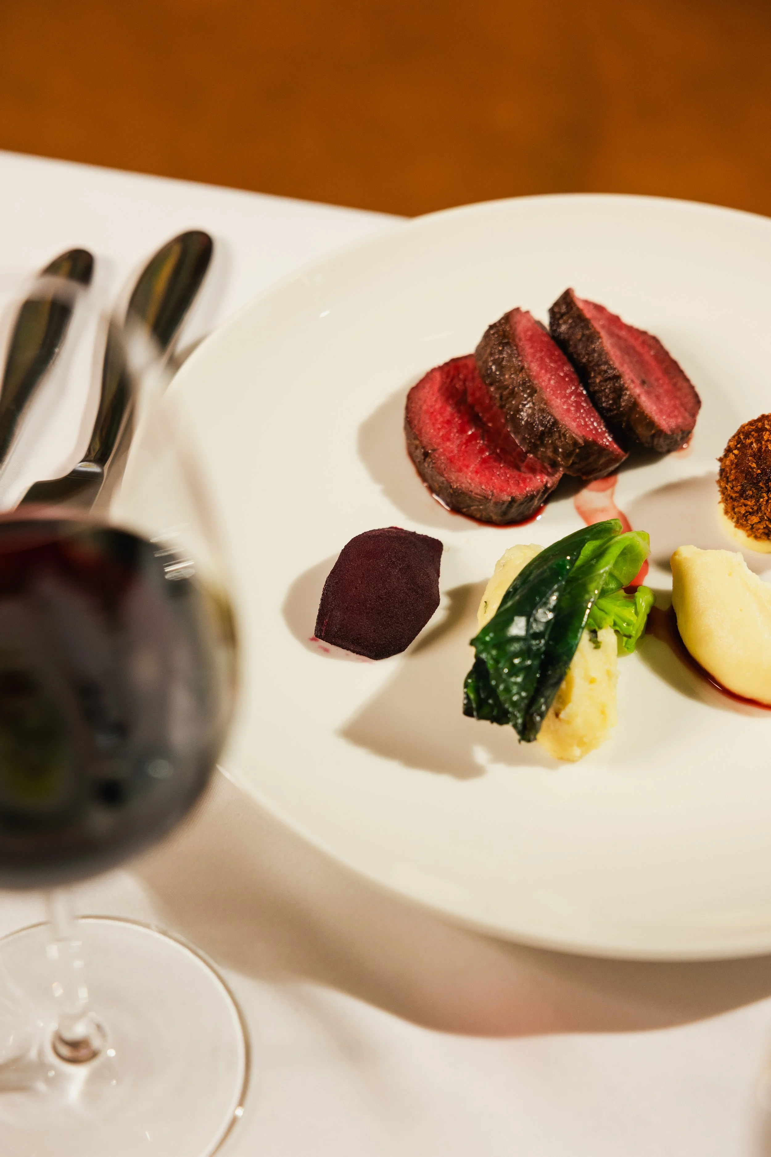 Slices of medium-rare steak on a white plate with vegetables, silverware, and a glass of red wine on a table.