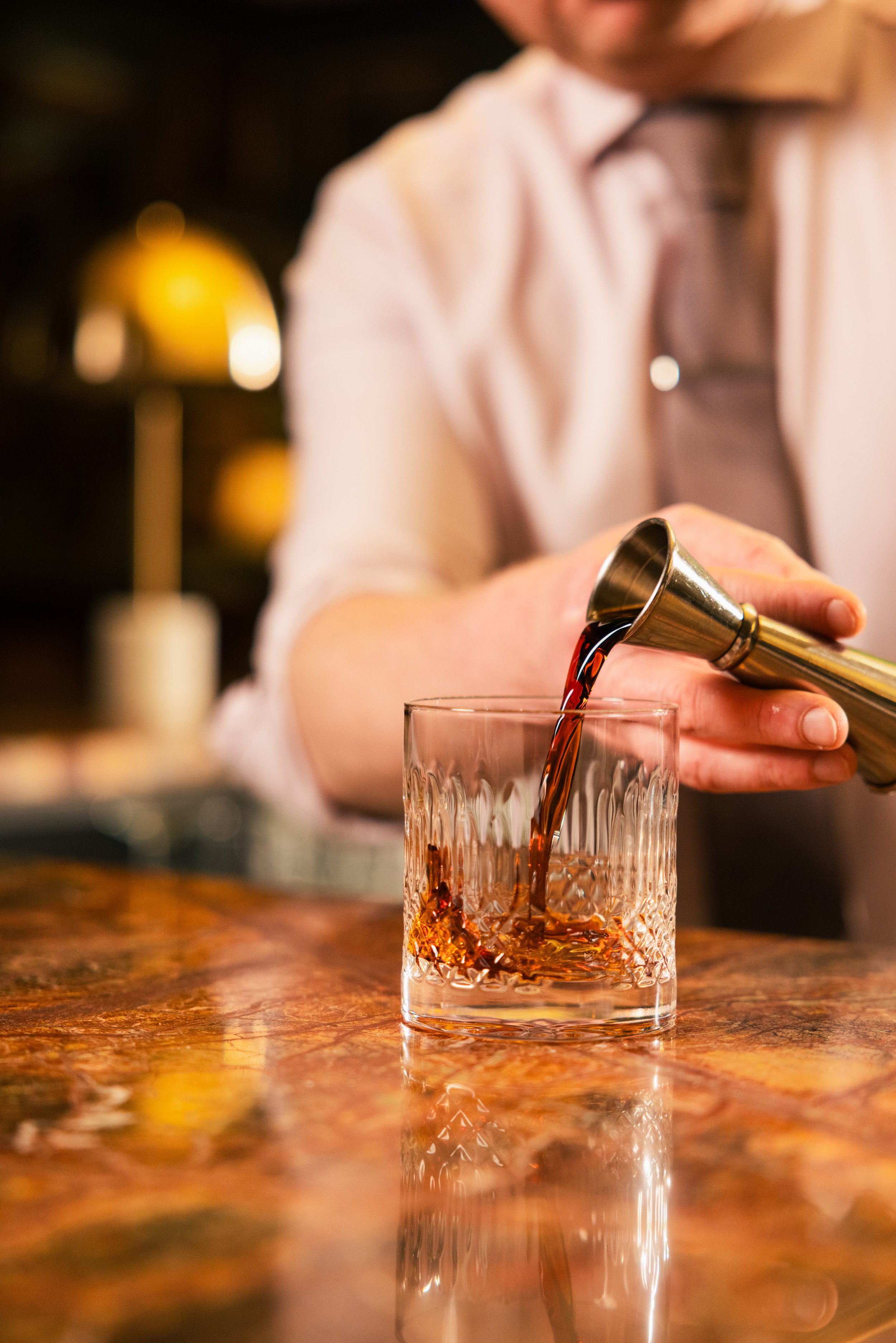 Person pouring a dark beverage into a crystal glass on a wooden table with a blurred background.