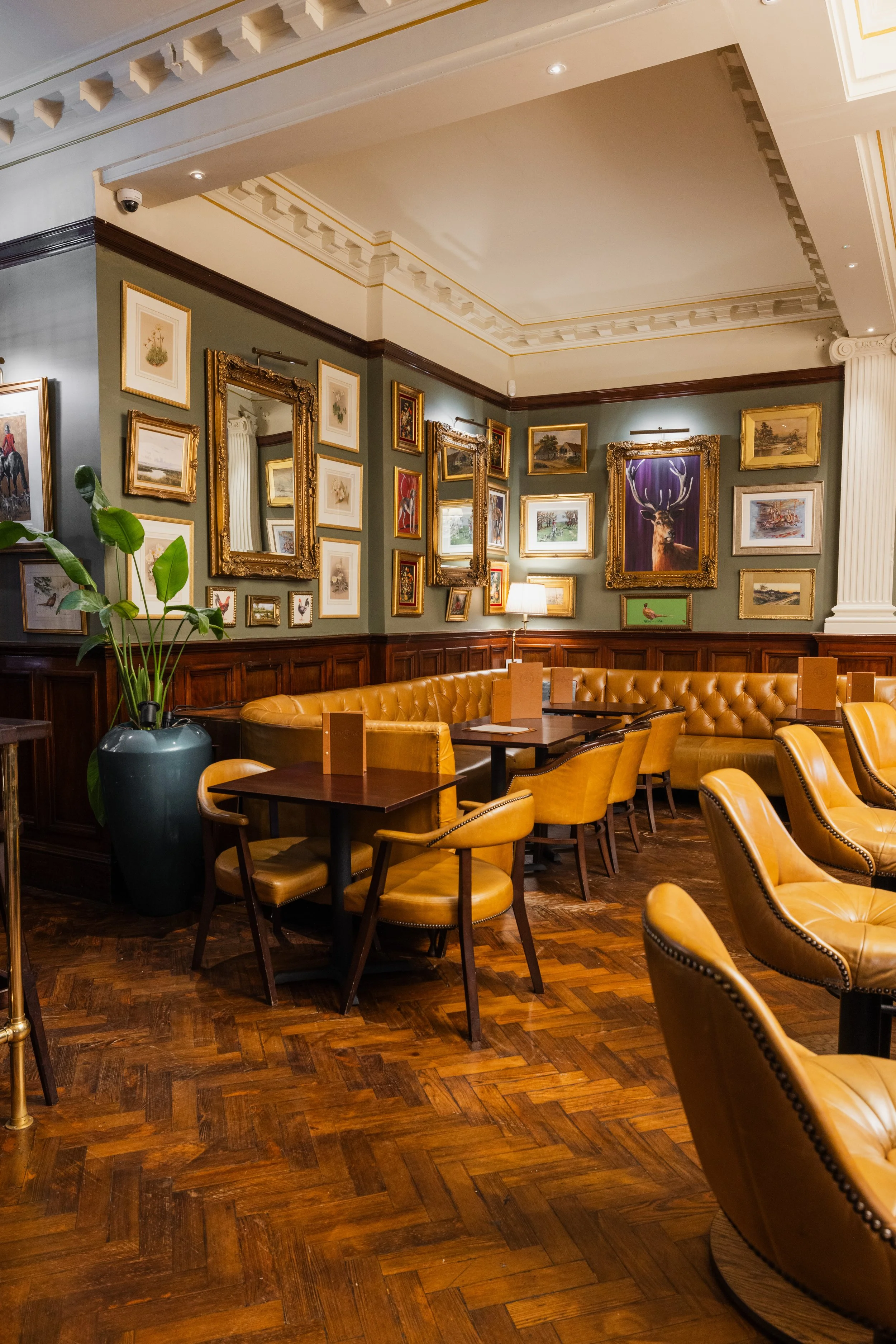 A cozy interior corner of a restaurant or cafe with a green painted wall adorned with various framed paintings and pictures, including landscapes, animals, and floral artwork. There are two ornate gold-framed mirrors. A wooden table surrounded by mus