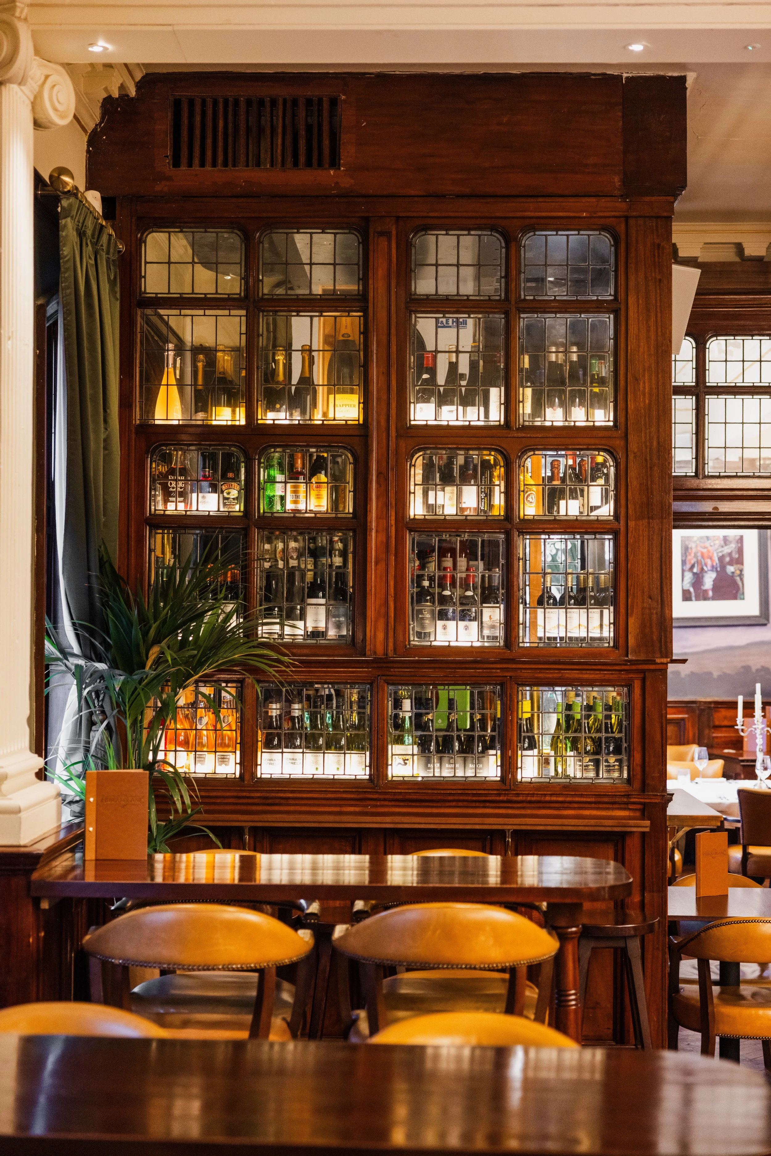 A wooden bar cabinet with glass windows displaying bottles of alcohol inside, set in a cozy restaurant with wooden furniture and yellow chairs, with decorative columns and green plants.
