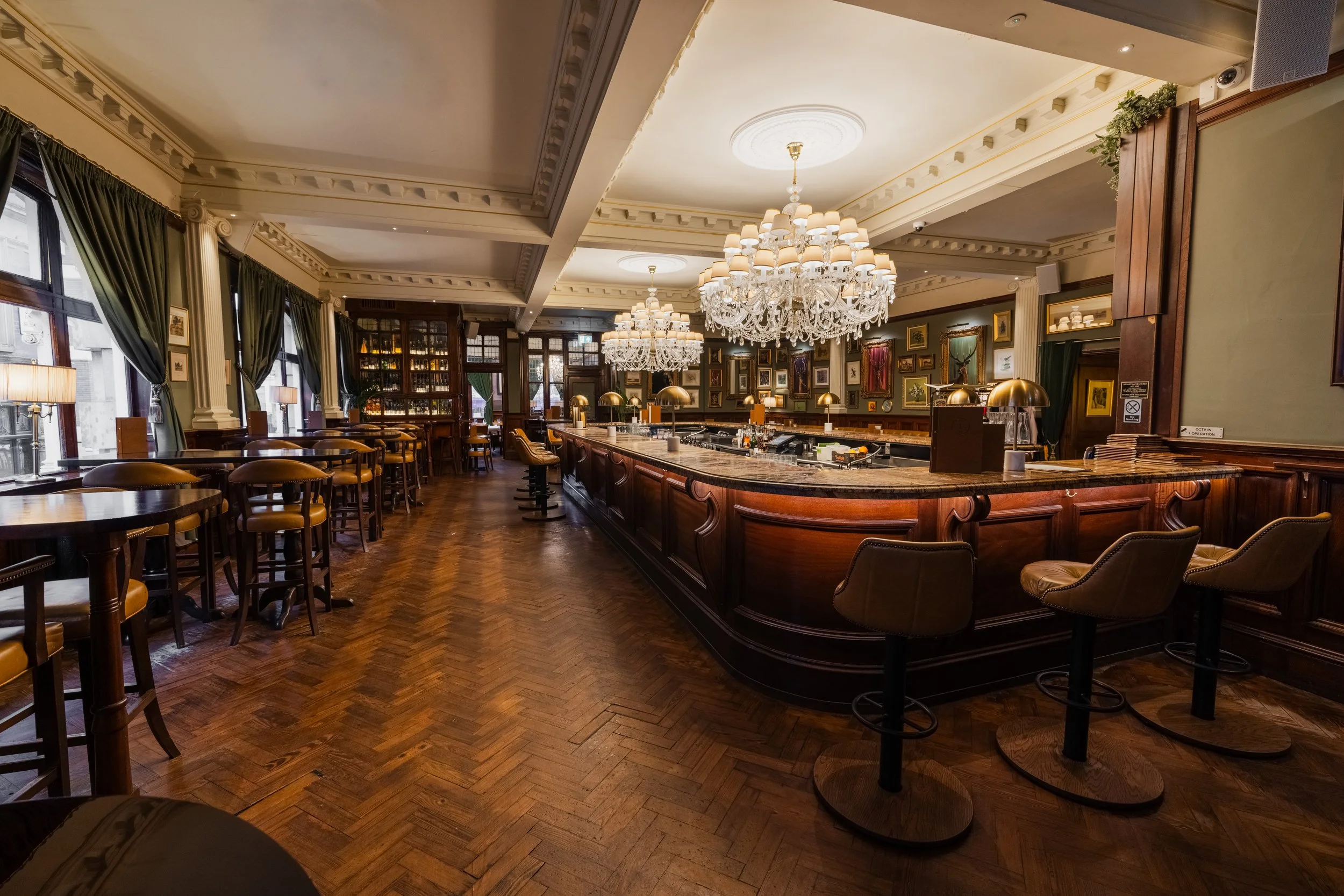 Elegant bar interior with chandeliers, wooden bar counter, barstools, large windows, dark green curtains, and framed artwork on the walls.