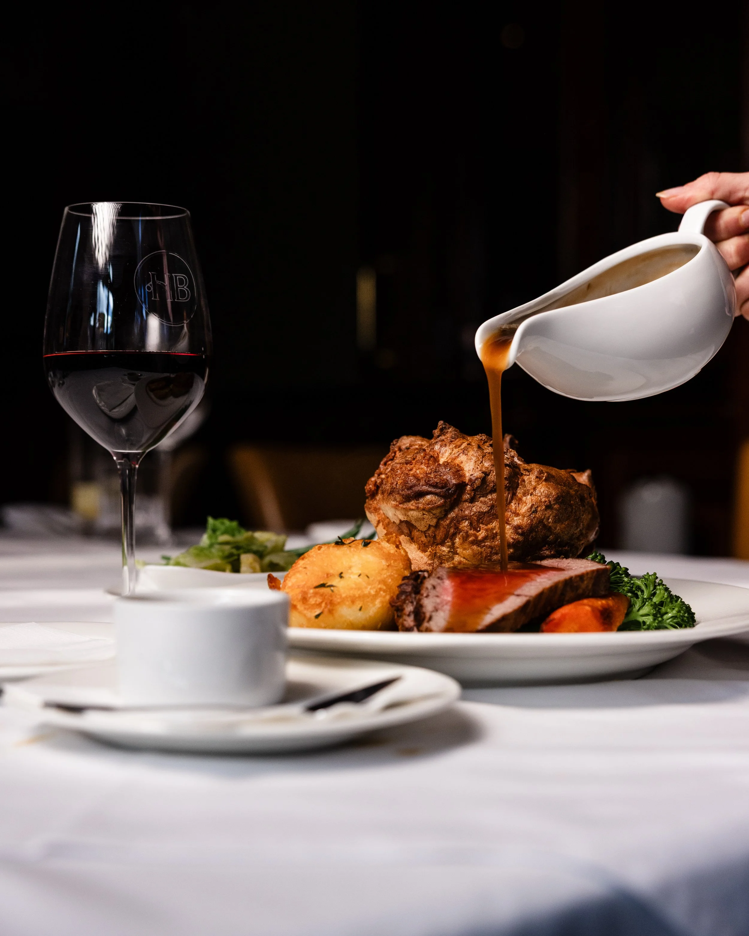 A glass of red wine and a plate with a roasted piece of meat, potatoes, vegetables, and sauce on a white table. Someone is pouring sauce over the meat from a white gravy boat.