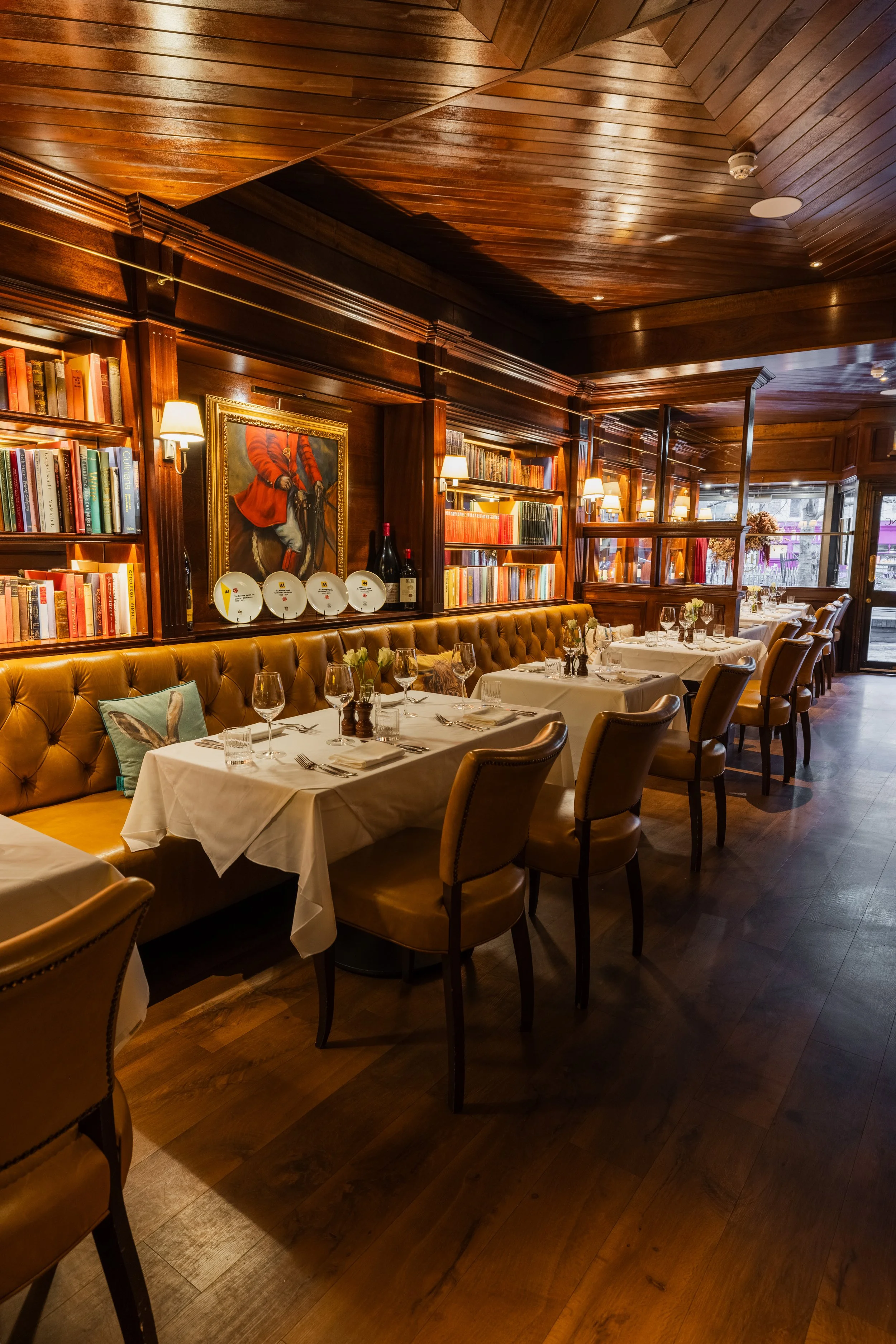 Elegant restaurant interior with wood-paneled walls and ceiling, bookshelves, framed artwork, wooden furniture, and table settings with wine glasses and tableware.