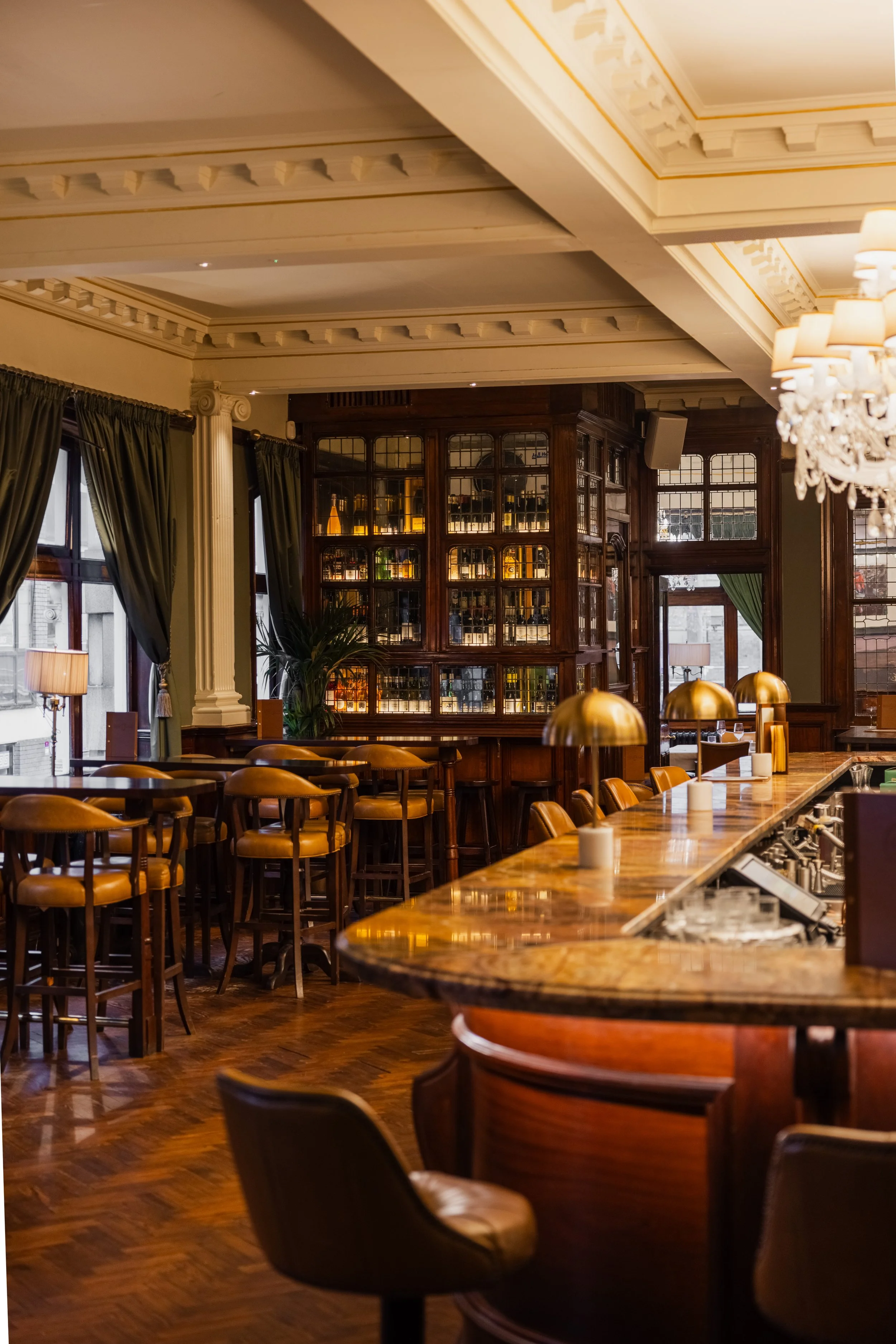 Interior of an elegant restaurant or bar with wooden furniture, large windows with green curtains, a marble-topped bar counter, and a wooden cabinet filled with bottles of alcohol.