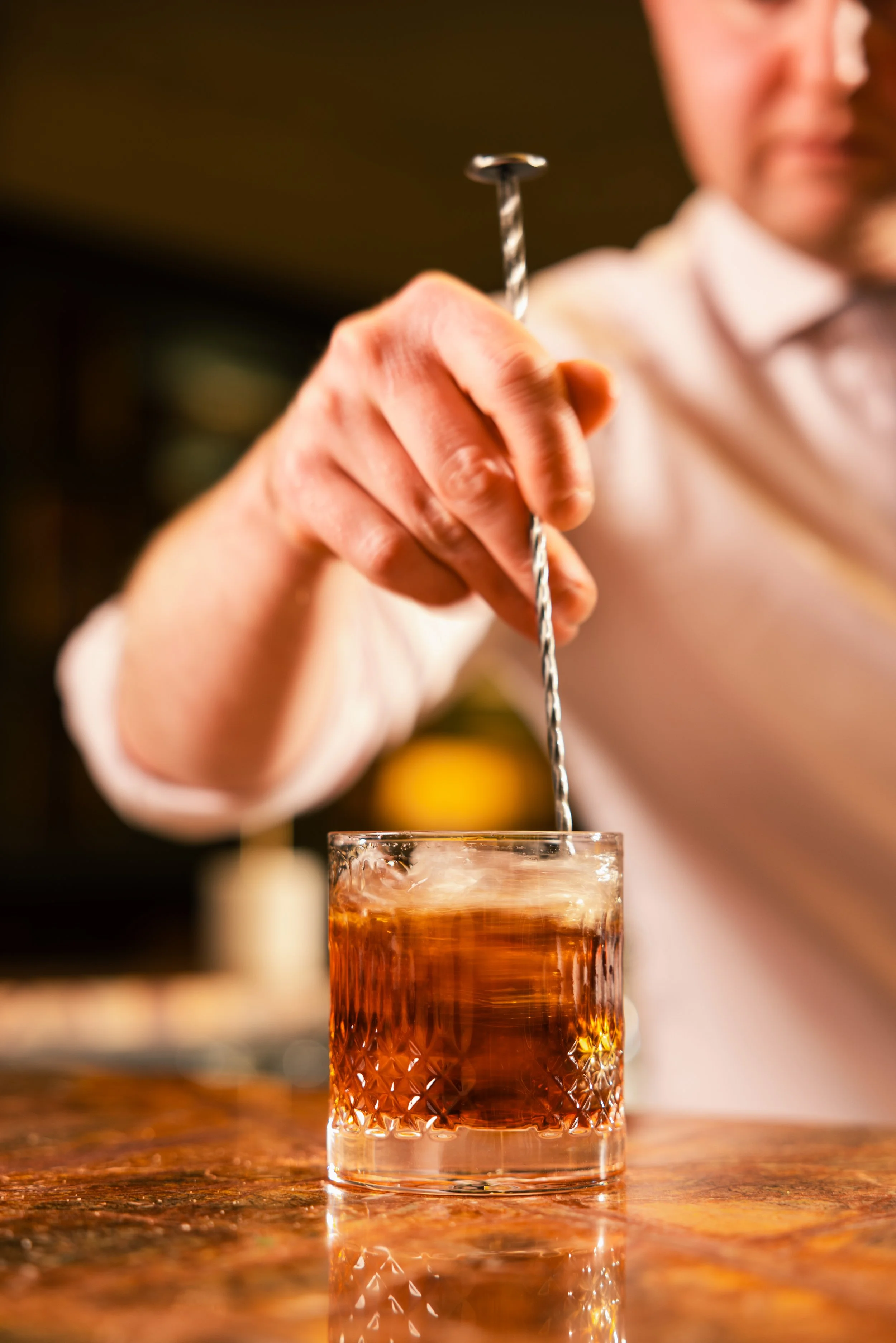 A person holding a long cocktail stir stick over a glass filled with amber-colored drink, with a blurred background.