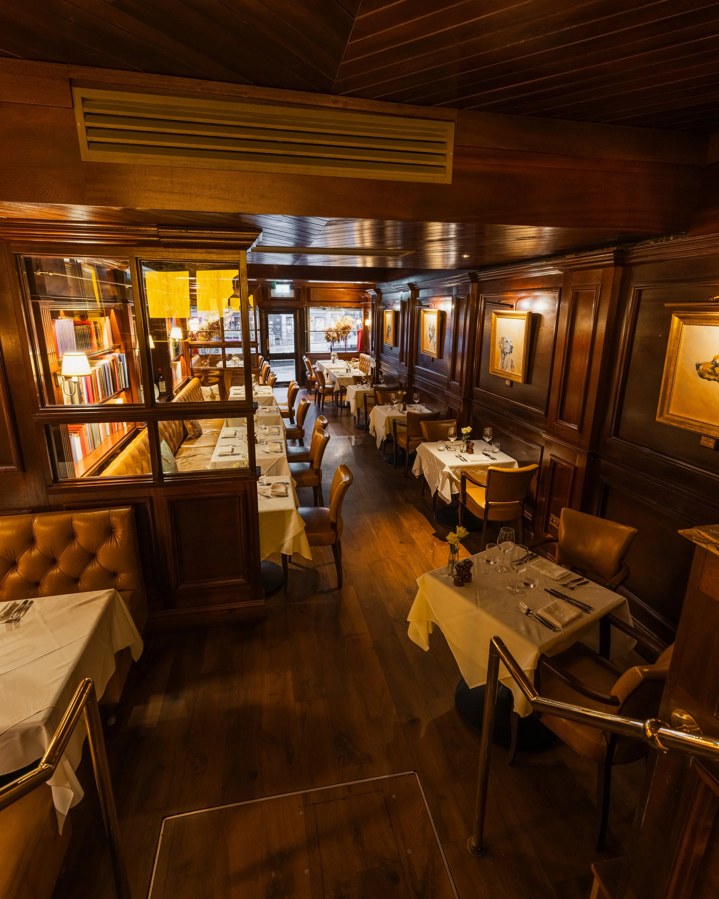 A cozy, upscale restaurant with white tablecloths, neatly set tables, and warm wooden interior walls adorned with framed artwork of dogs. The restaurant is illuminated by soft lighting, and natural light streams in through large windows at the back.