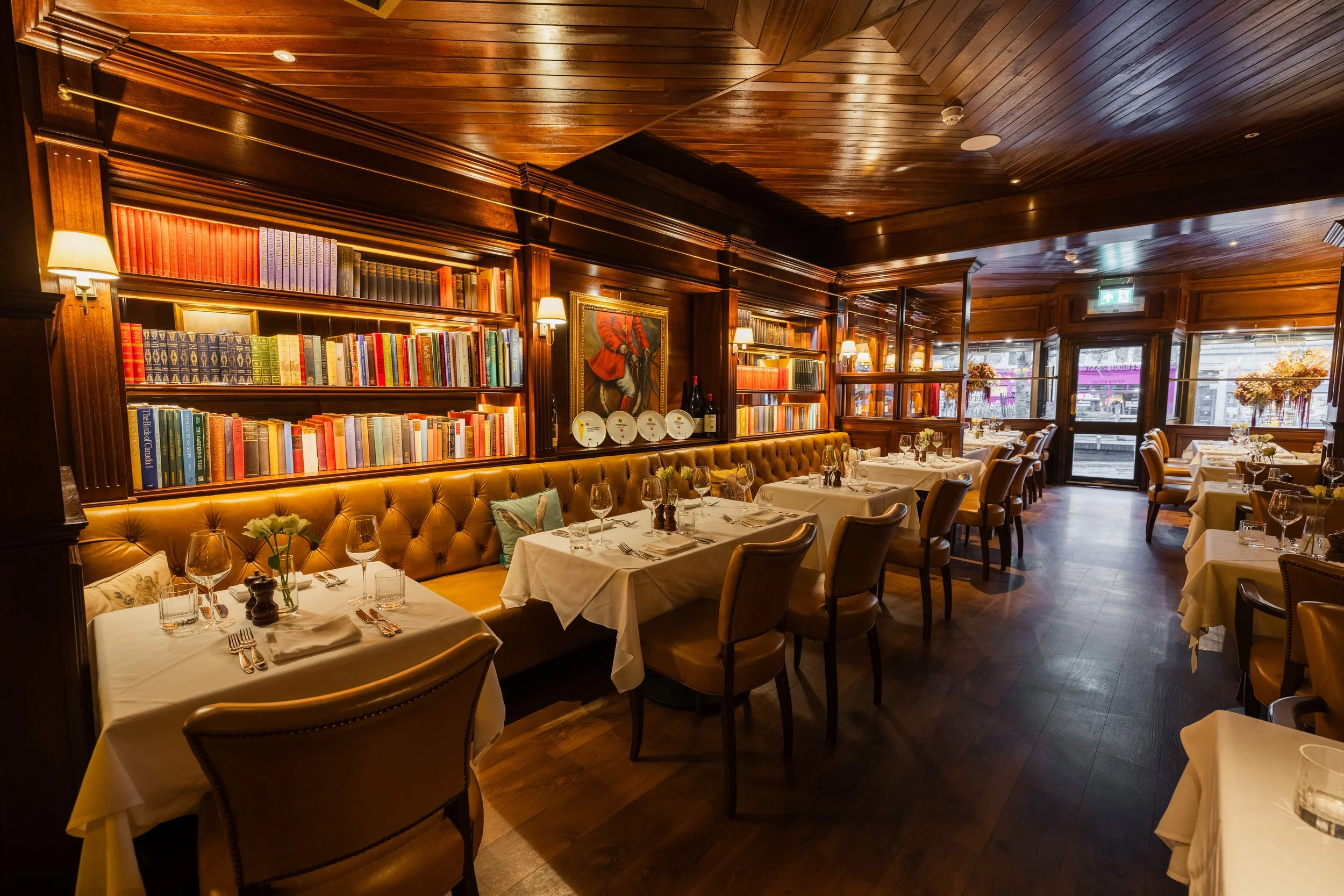 Inside a cozy restaurant with wooden walls, bookshelves, tables set with wine glasses, and white tablecloths. There are large windows with flowers outside and warm lighting.