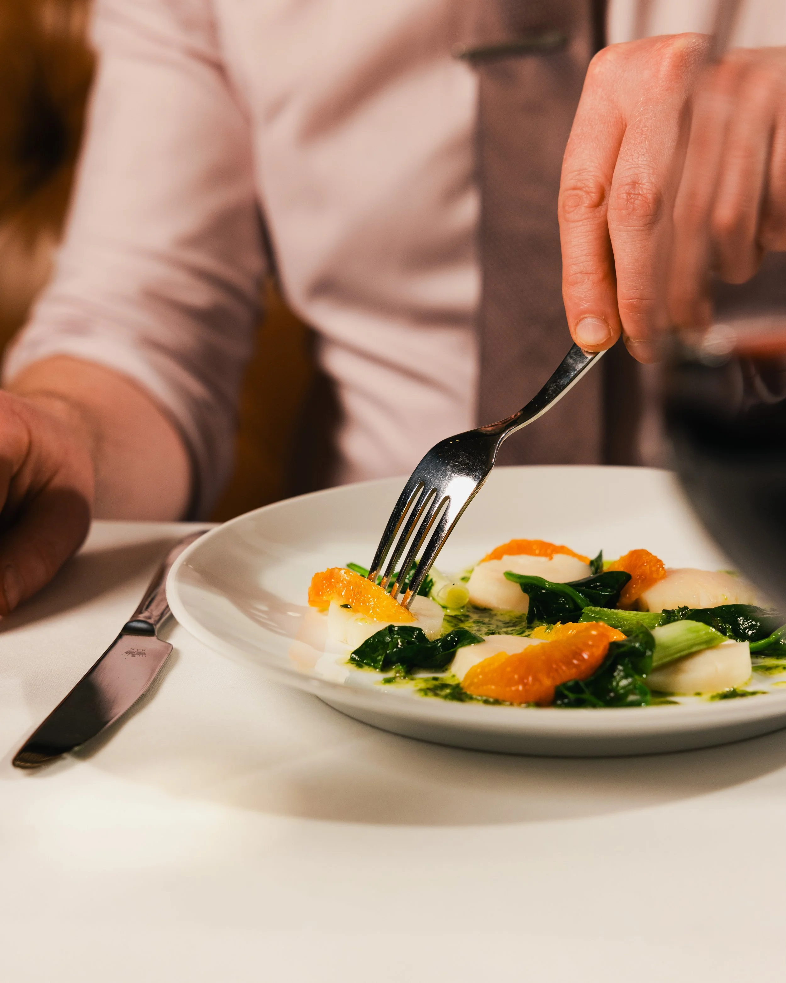 Close-up of a person using a fork to eat a dish with white fish fillet, orange slices, and greens on a white plate.