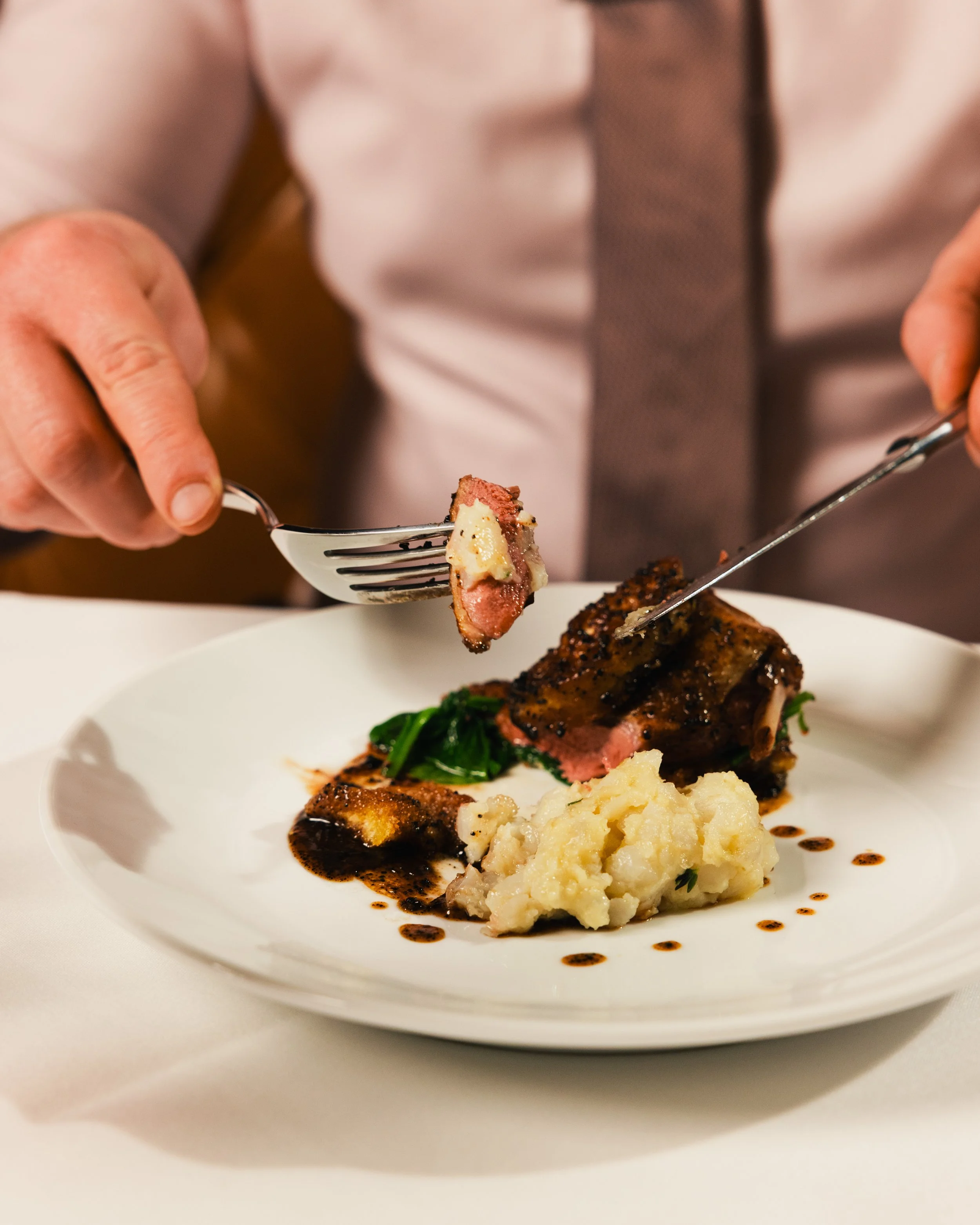 Close-up of a gourmet dish on a white plate, featuring a piece of grilled meat with mashed potatoes and greens, garnished with a dark sauce.