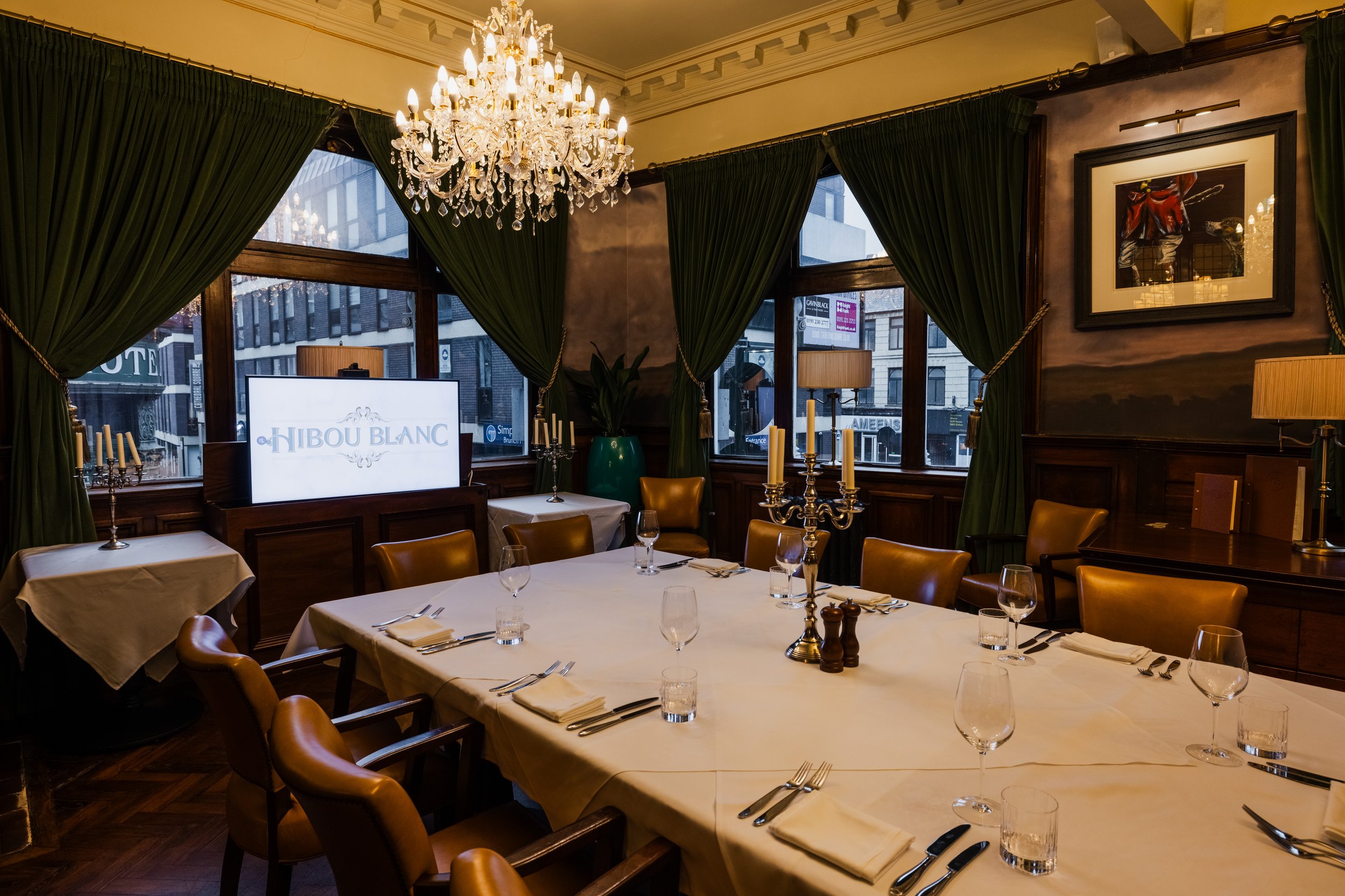 Elegant dining room with a chandelier, dark green curtains, framed artwork, and a table set with white tablecloths, glassware, and silverware.