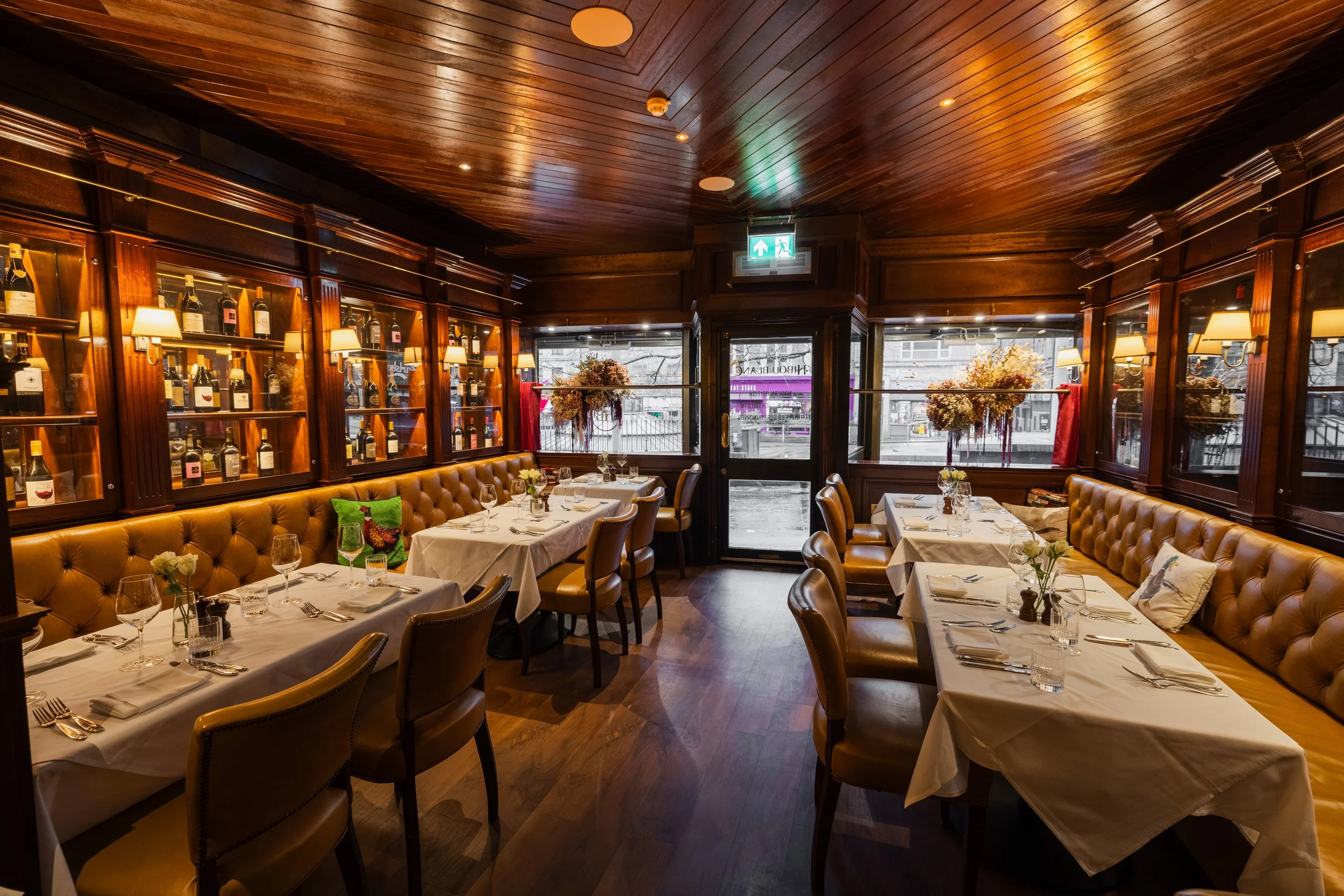 Interior of a restaurant with wooden ceiling and walls, decorated with shelves of wine bottles, floral arrangements, and tables set with white tablecloths, glasses, and silverware.