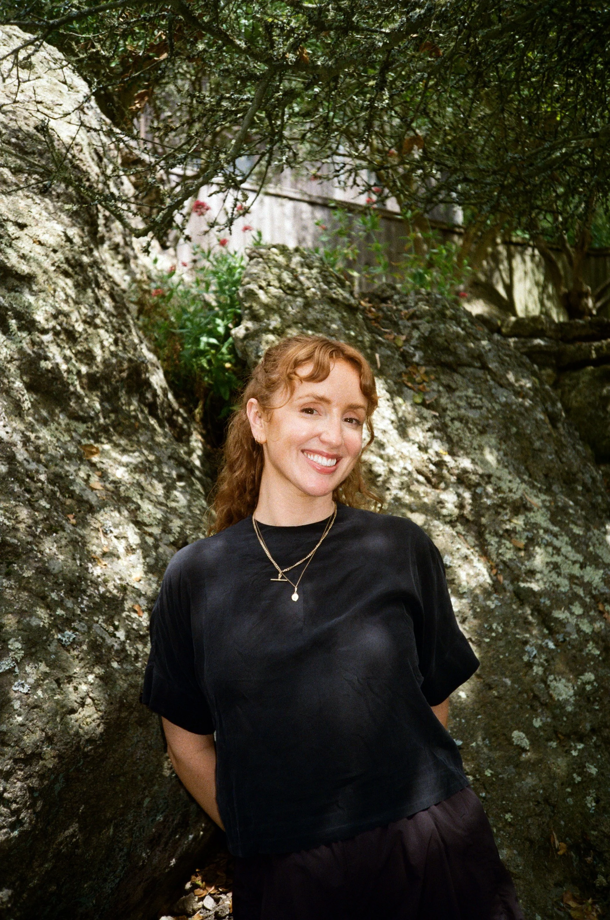A woman with reddish hair, smiling, standing outdoors in front of a natural rocky background with trees and plants.