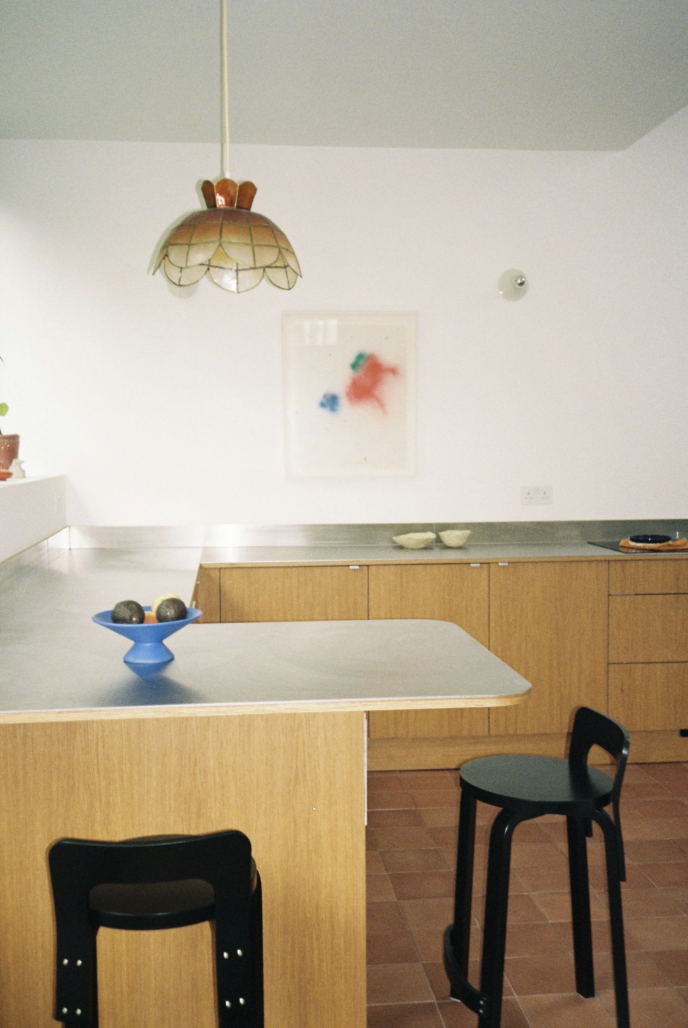 A kitchen with a hanging stained glass lamp, a white wall with abstract red and blue artwork, and a wooden countertop with bowls and a black chair.