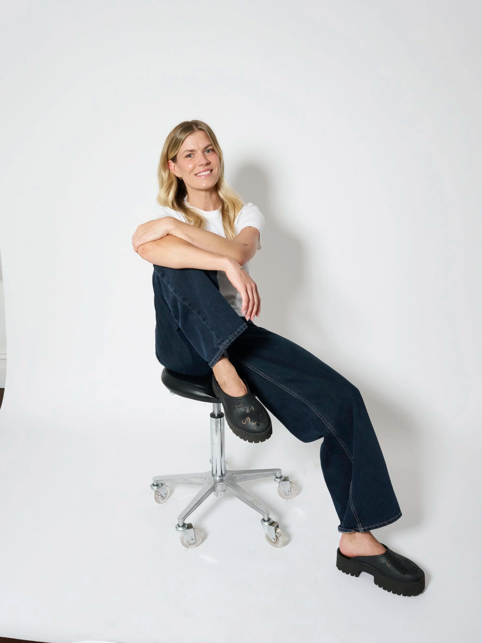 A woman sitting on a stool with her arms crossed, wearing a white t-shirt, dark jeans, black platform shoes, with a neutral background.