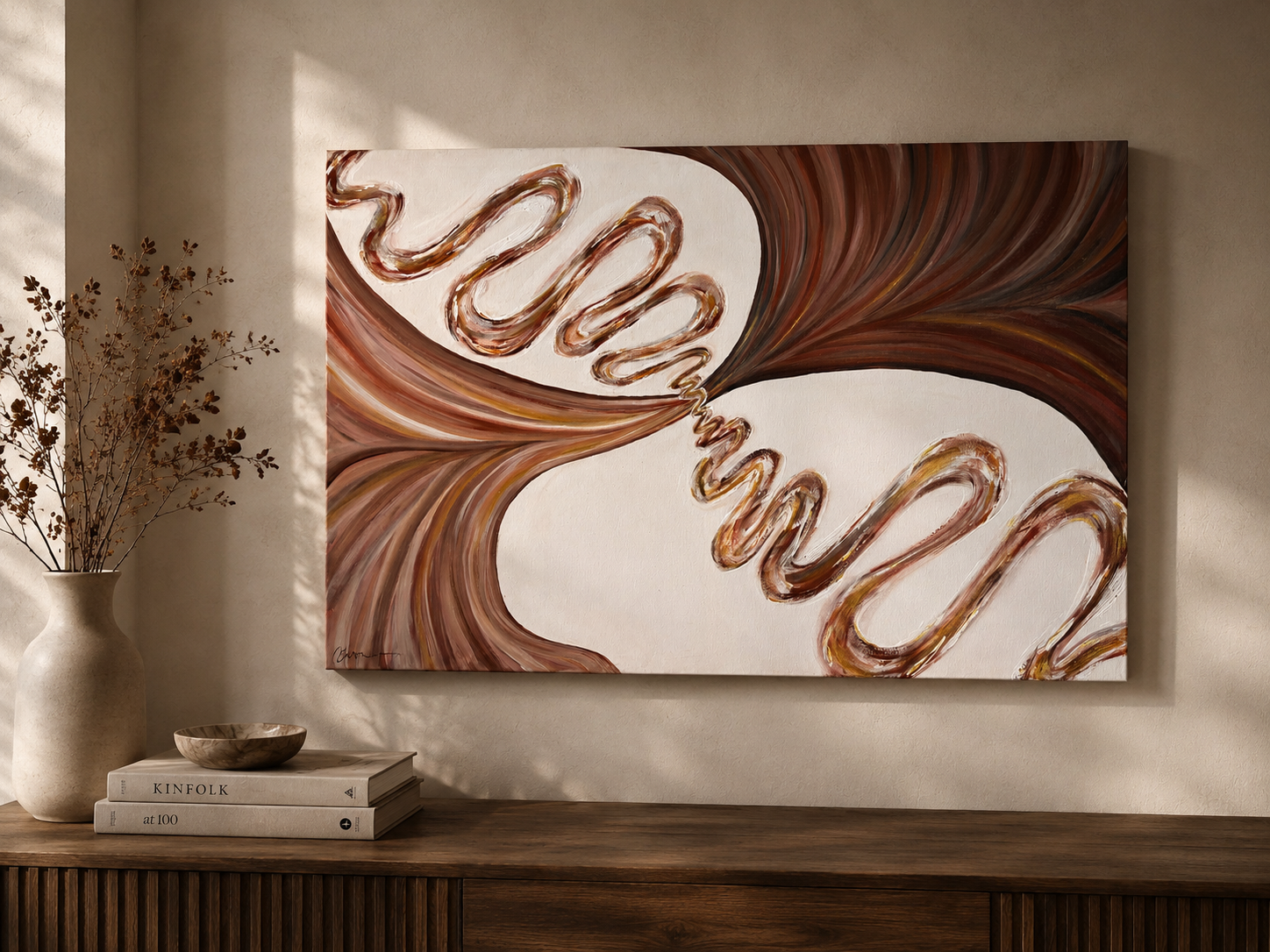 Abstract painting with swirling brown and white patterns hanging on a beige wall, with a white vase with branches and a stack of books on a wooden surface below.