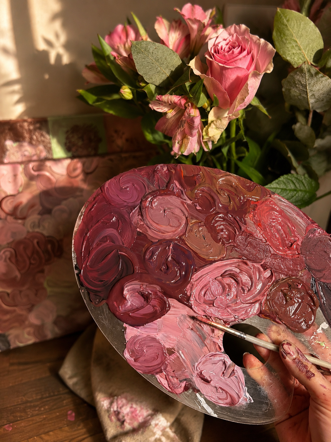 An artist's hand holding a palette with pink and red oil paints, with a bouquet of pink roses and greenery in the background.