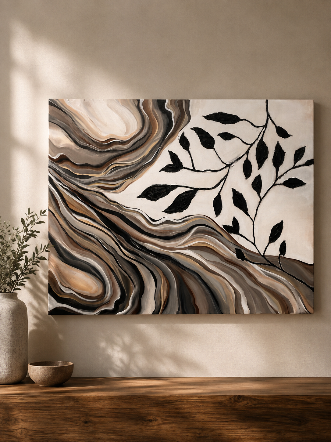Abstract wall art with black leaves and swirling patterns in neutral tones, hanging on a beige wall with a wooden surface below, decorated with a potted plant and bowls.
