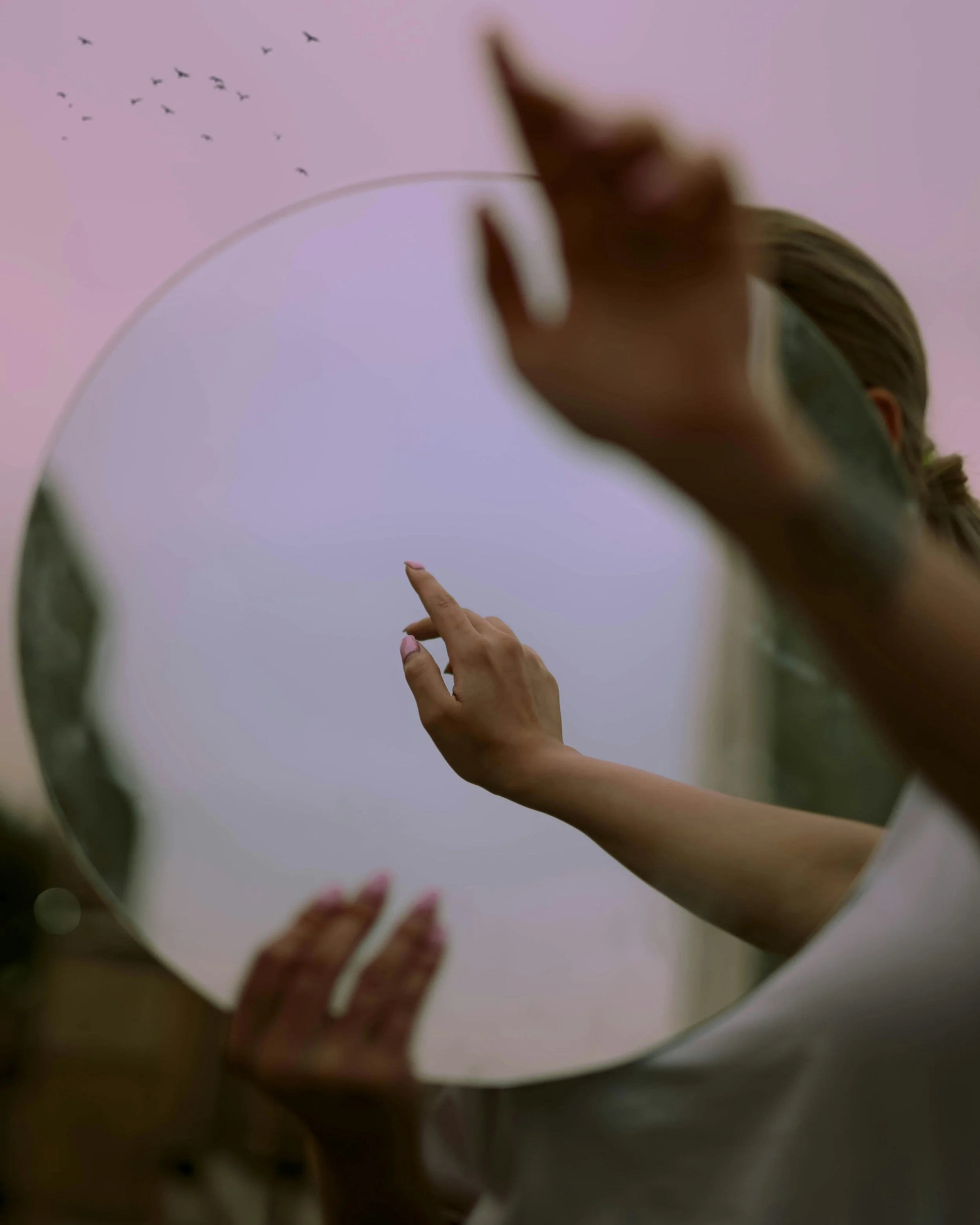 A person's hand and finger pointing to a mirror. The mirror reflects part of a person's face and a group of birds flying in a pink-purple sky.