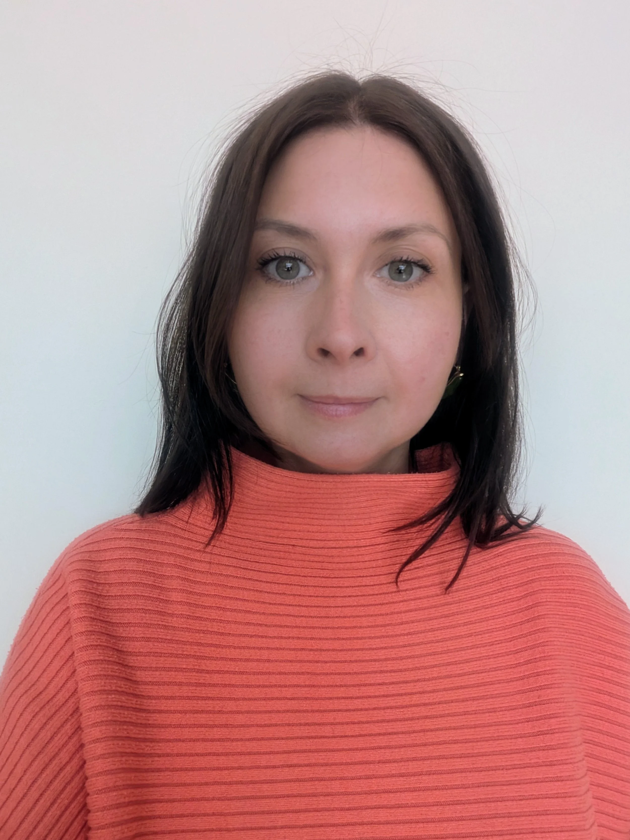A woman with long dark hair, green eyes, and light-medium skin, wearing a coral-colored ribbed sweater, stands against a plain white wall.