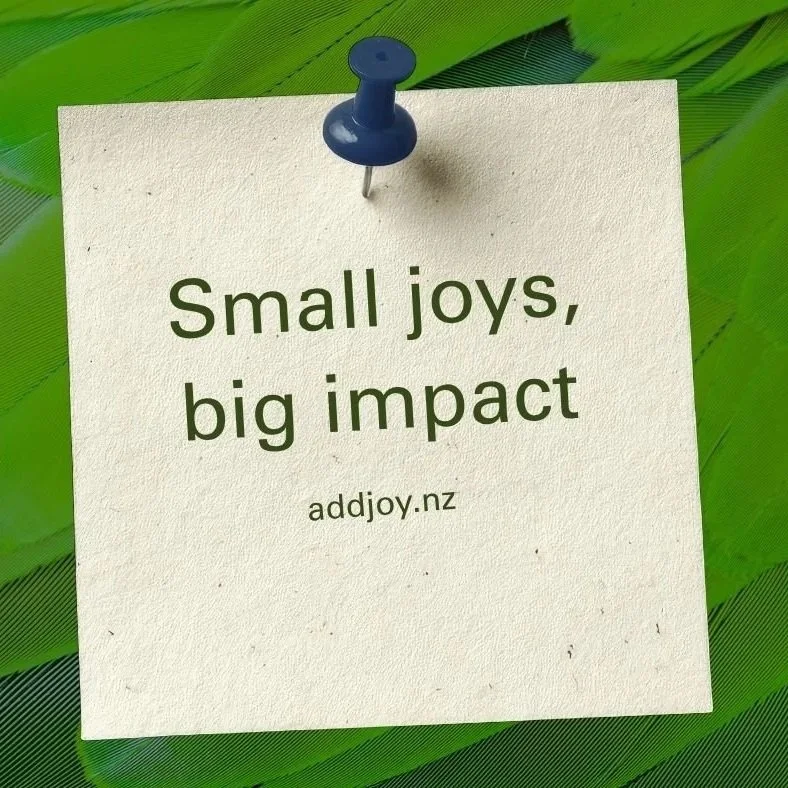 A white note pinned with a blue pushpin on green leaves, with the text "Small joys, big impact" and the website www.addjoy.nz