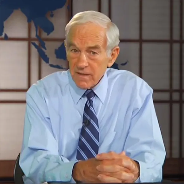 Older man with white hair in a light blue shirt and striped tie speaking in front of a world map.