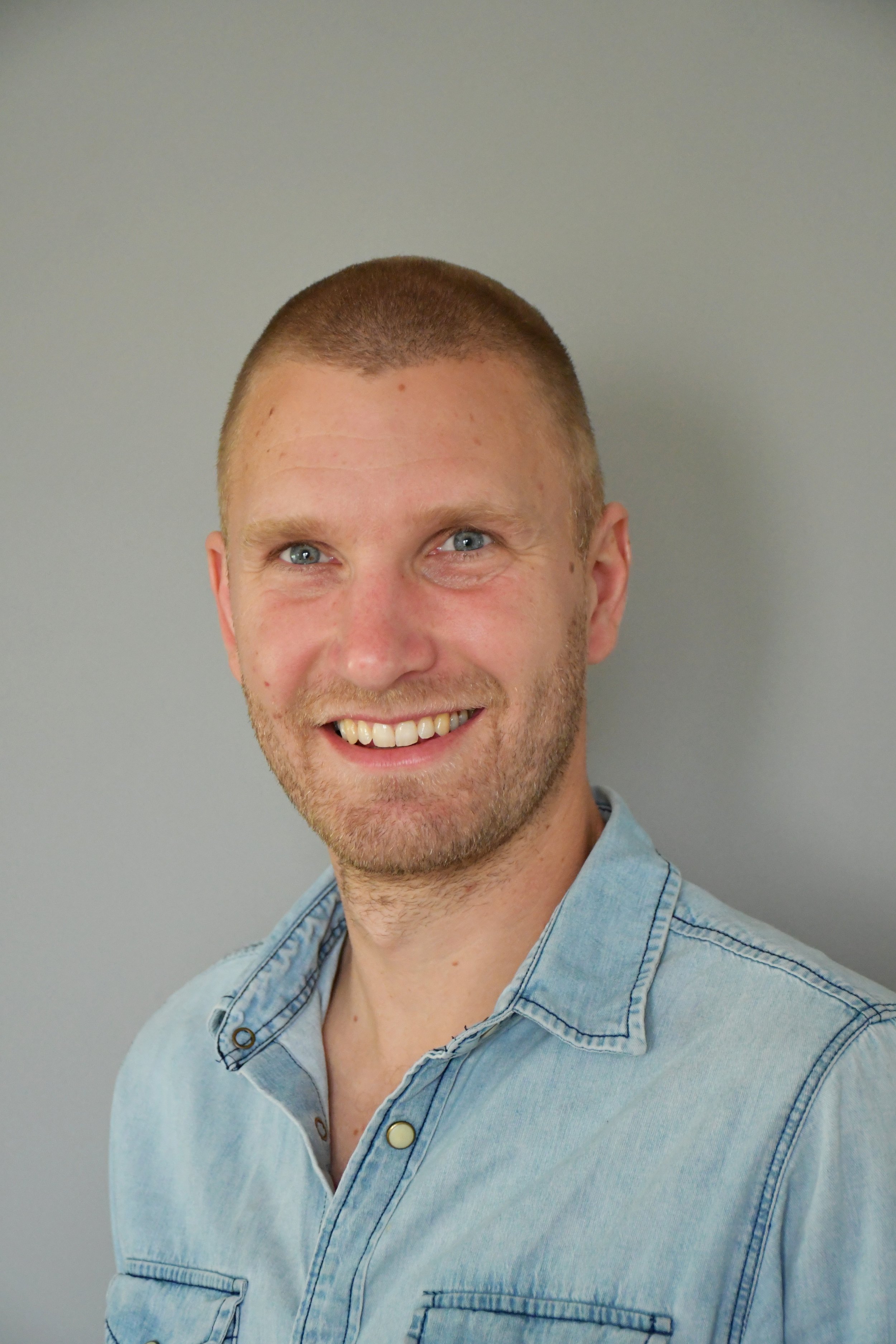 Junger Mann mit kurzen, hellbraunen Haaren, blauäugig, im Jeanshemd, lächelnd vor grauem Hintergrund.