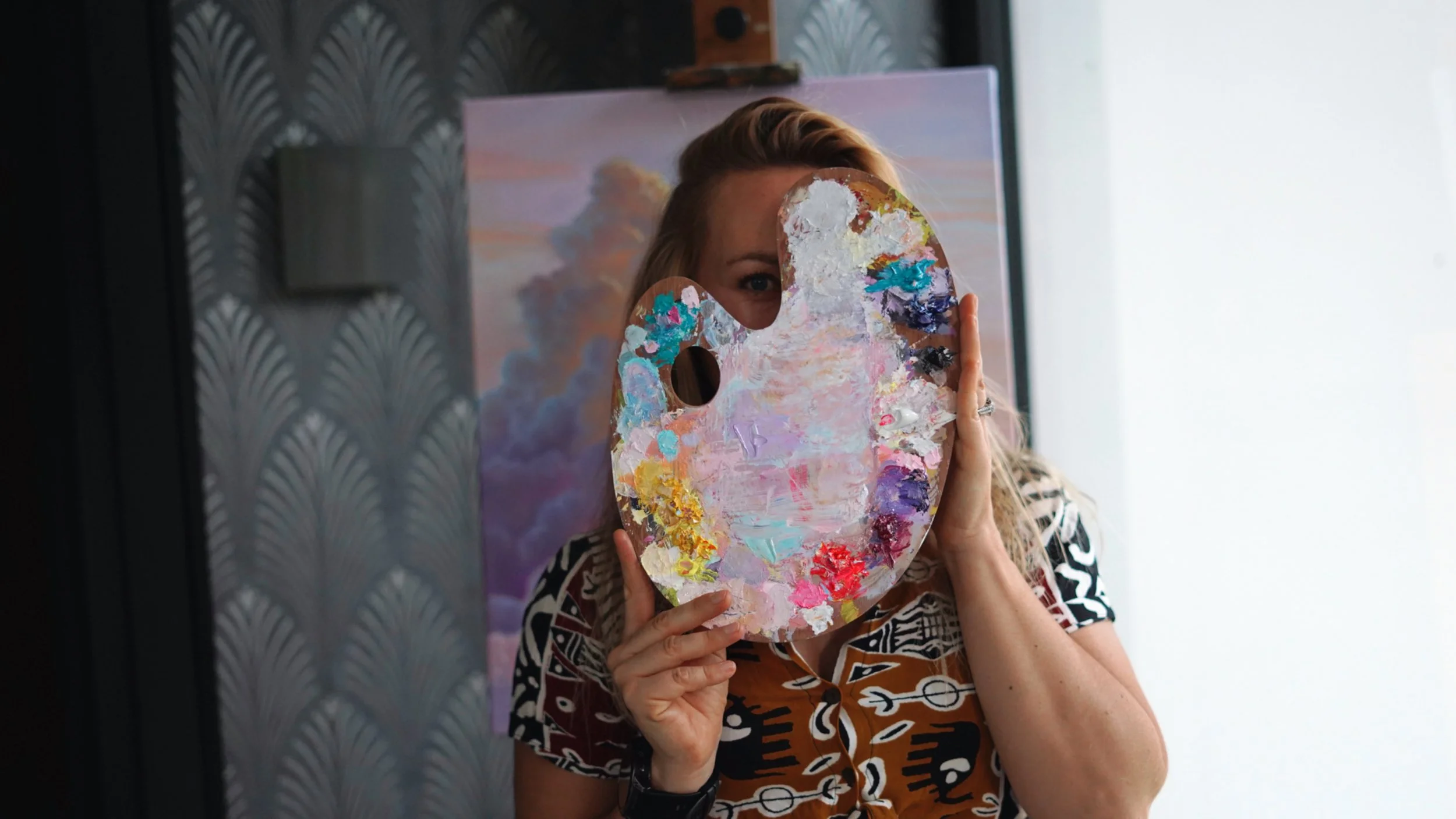 A woman holding a painted artist's palette in front of her face, with part of her eye visible behind the palette. She is standing indoors near a window, with artwork and a patterned partition in the background.