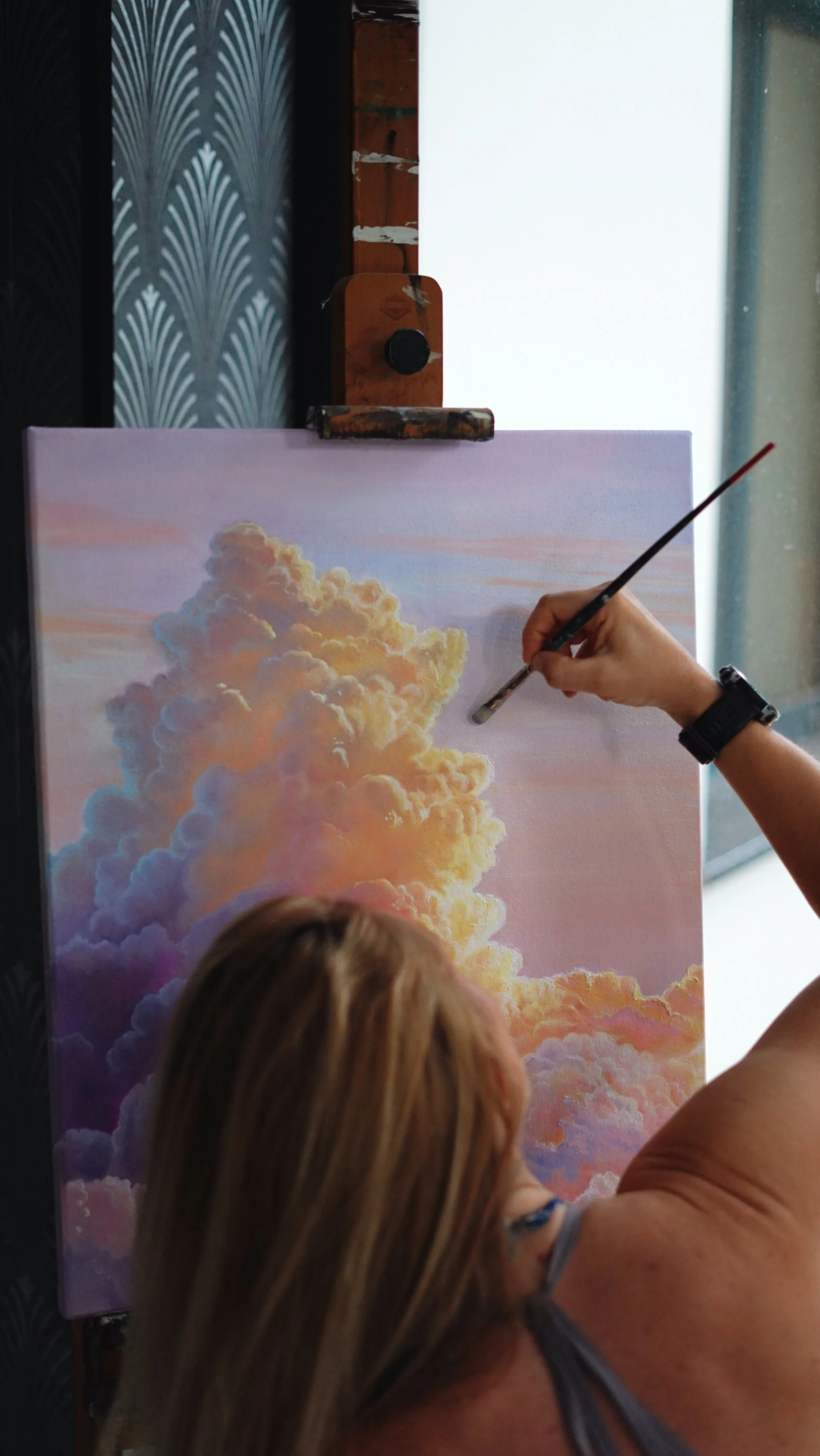 An artist is painting a sky scene with fluffy clouds on a large canvas, using a brush to add details.